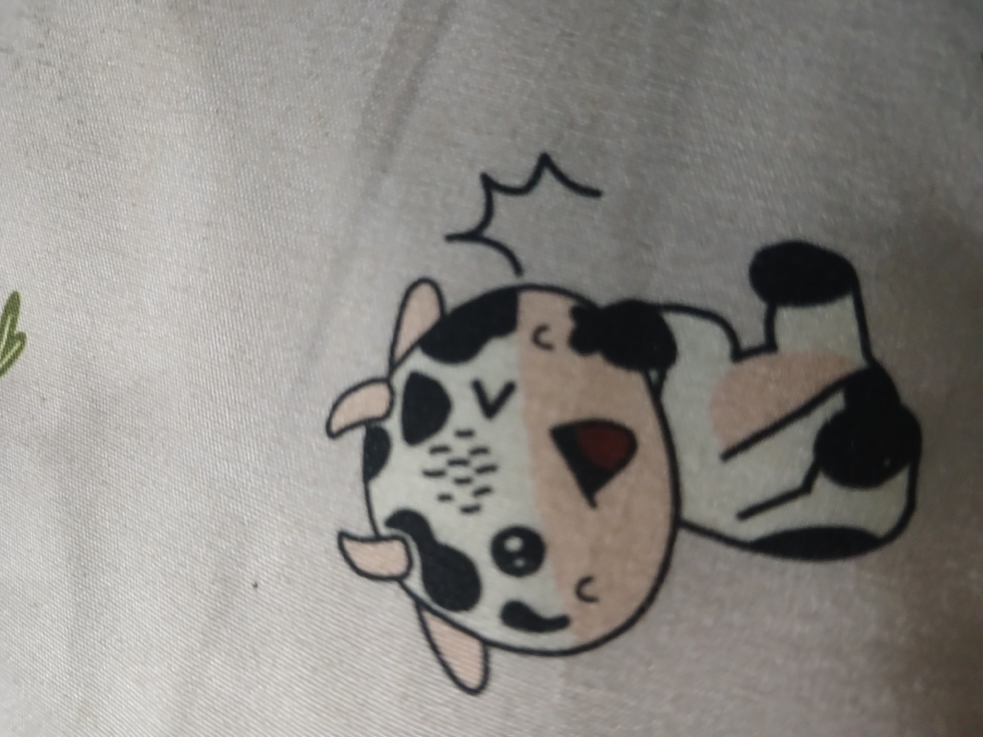 The image shows a close-up of a white textile with a printed cartoon cow. The cow is black and white with pink accents on its ears and cheeks. It is sitting down and has its left hand over its mouth, suggesting it is laughing or trying to suppress a giggle. There are black "sparkle" lines near its head, indicating excitement or surprise. The cow has a black outline and appears to be part of a larger pattern or design on the fabric. The environment is a plain, textured surface, likely a piece of cloth or bedding. There are no indications of the city of Randuagung, Indonesia, in the image.