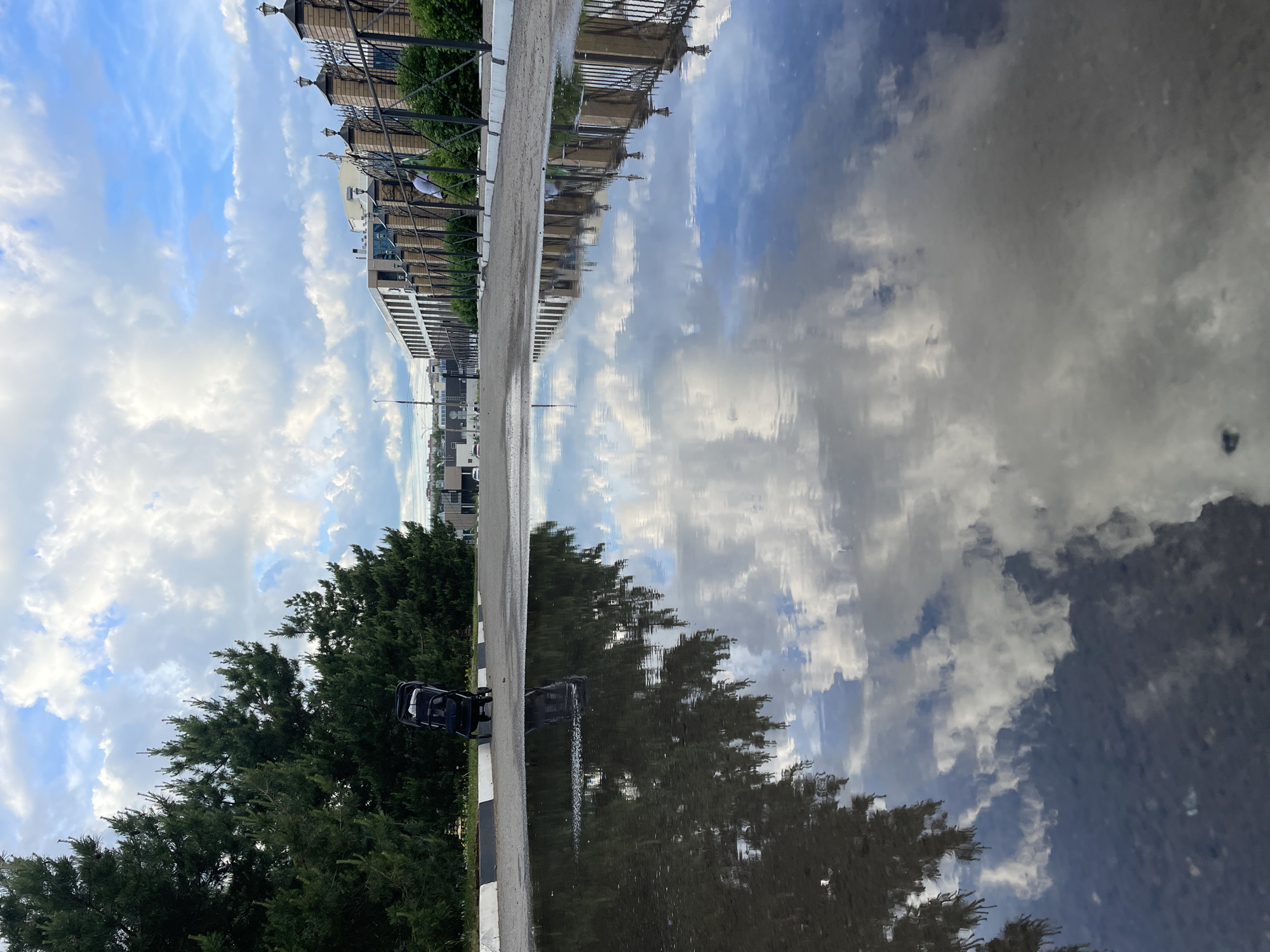 A street scene in Izhevsk, Russia. The image is rotated to the left. A puddle reflects the sky, trees, and buildings, creating a mirrored effect. On the left of the puddle, there is a street. A black stroller is positioned next to evergreen trees. Along the opposite side of the street, there are apartment buildings. The sky is filled with clouds.