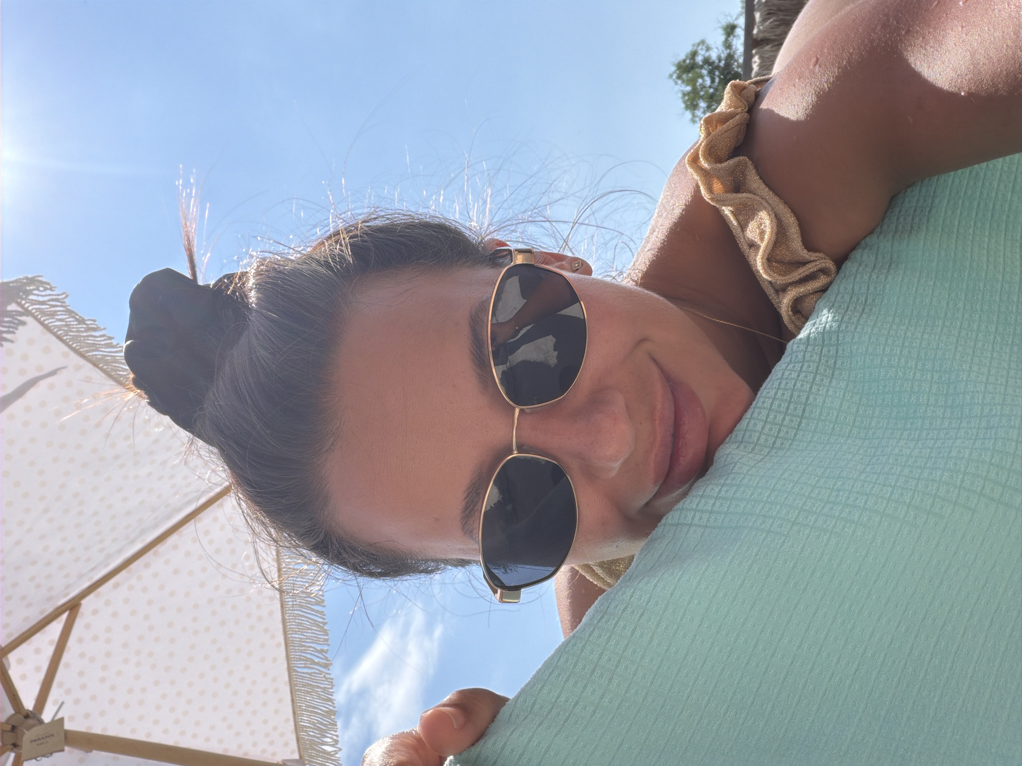 A person is pictured outdoors, under a bright sun and blue sky, partially covered by a light-colored, polka-dotted umbrella with fringed edges. The individual is wearing gold-rimmed sunglasses with dark lenses and a gold, ruffled bikini top. They are leaning on a textured, light blue towel or blanket. The scene appears to be a sunny day, possibly at a beach or resort in Burgas, Bulgaria, indicated by the presence of the umbrella and the overall summery atmosphere.