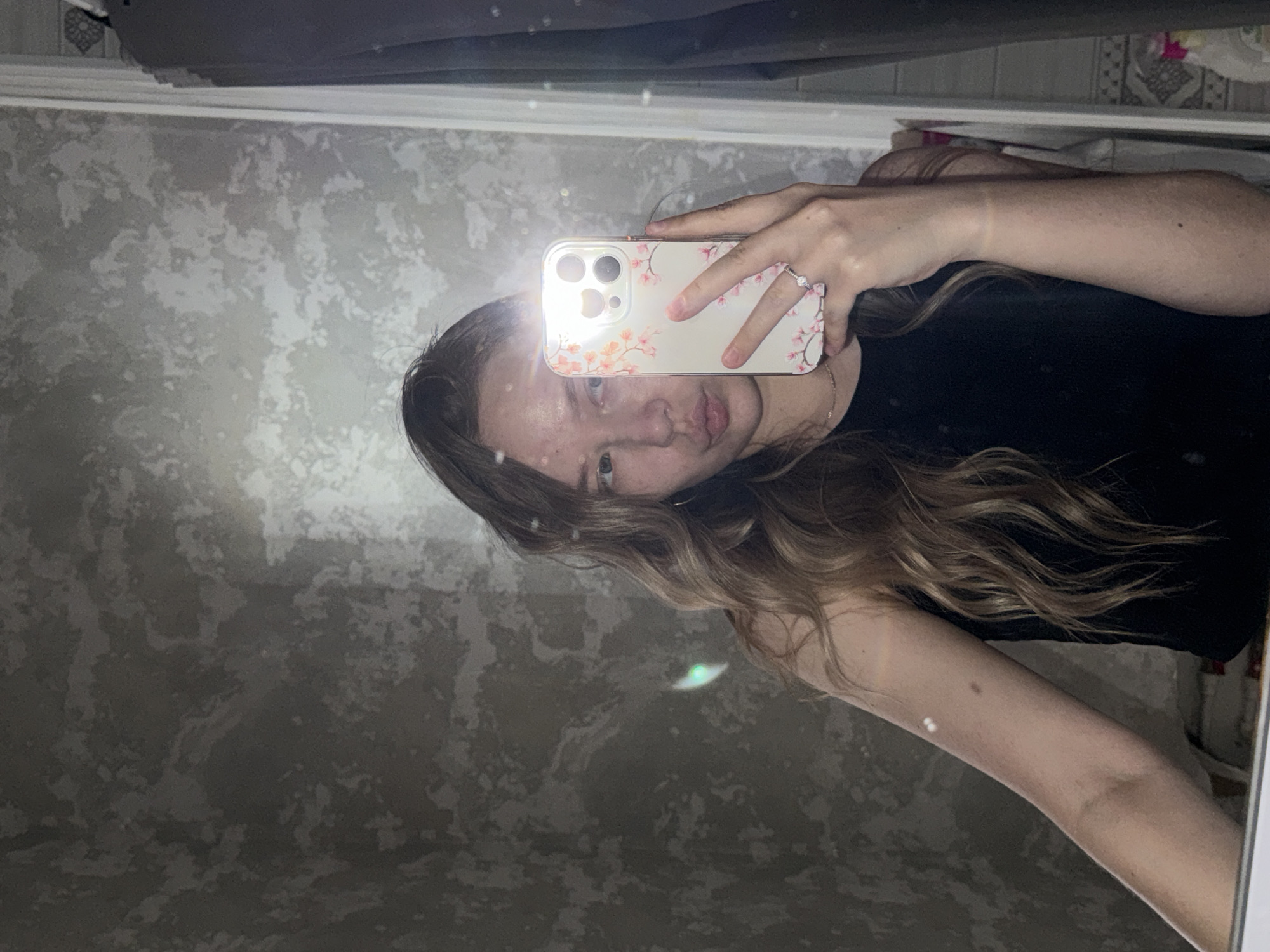 This is a mirror selfie taken indoors, likely in a bathroom or bedroom, given the textured wallpaper and the presence of a mirror. The main subject is a young person, likely female, with long, wavy, light brown hair. She is holding a smartphone in her right hand, which is angled to capture her reflection. The phone has a light-colored case with a floral pattern featuring pink cherry blossoms and green leaves. She is wearing a black top. Her left arm is visible, and she is wearing a silver ring with small stones on her left ring finger.
The lighting in the image suggests it is daytime, with a bright flash from the phone illuminating her face and creating some lens flare. Her facial expression is relaxed, with her lips slightly puckered. The background is a textured wallpaper with a marble-like pattern in shades of grey and white. The reflection also shows a portion of the ceiling and what appears to be a shelf or ledge above the mirror. The overall mood is casual and personal. No visible text is present in the image.