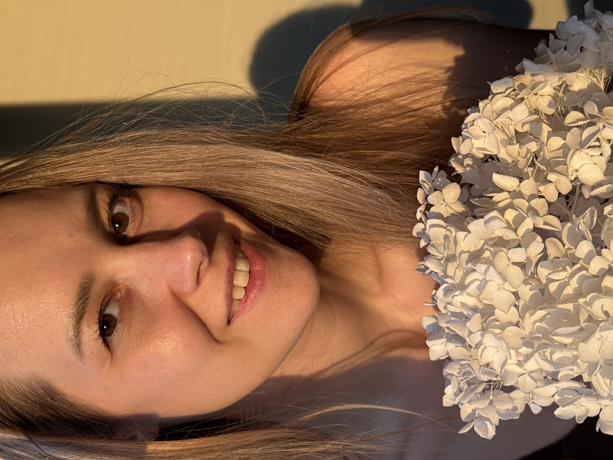 A young woman with light brown hair is lying down, smiling at the camera. She has light skin, brown eyes, and her head is turned slightly to the left. A cluster of white flowers rests near her right shoulder. A shadow falls across the top of her head. The background is a solid, light color, and the location is Chaikovsky, Russia.