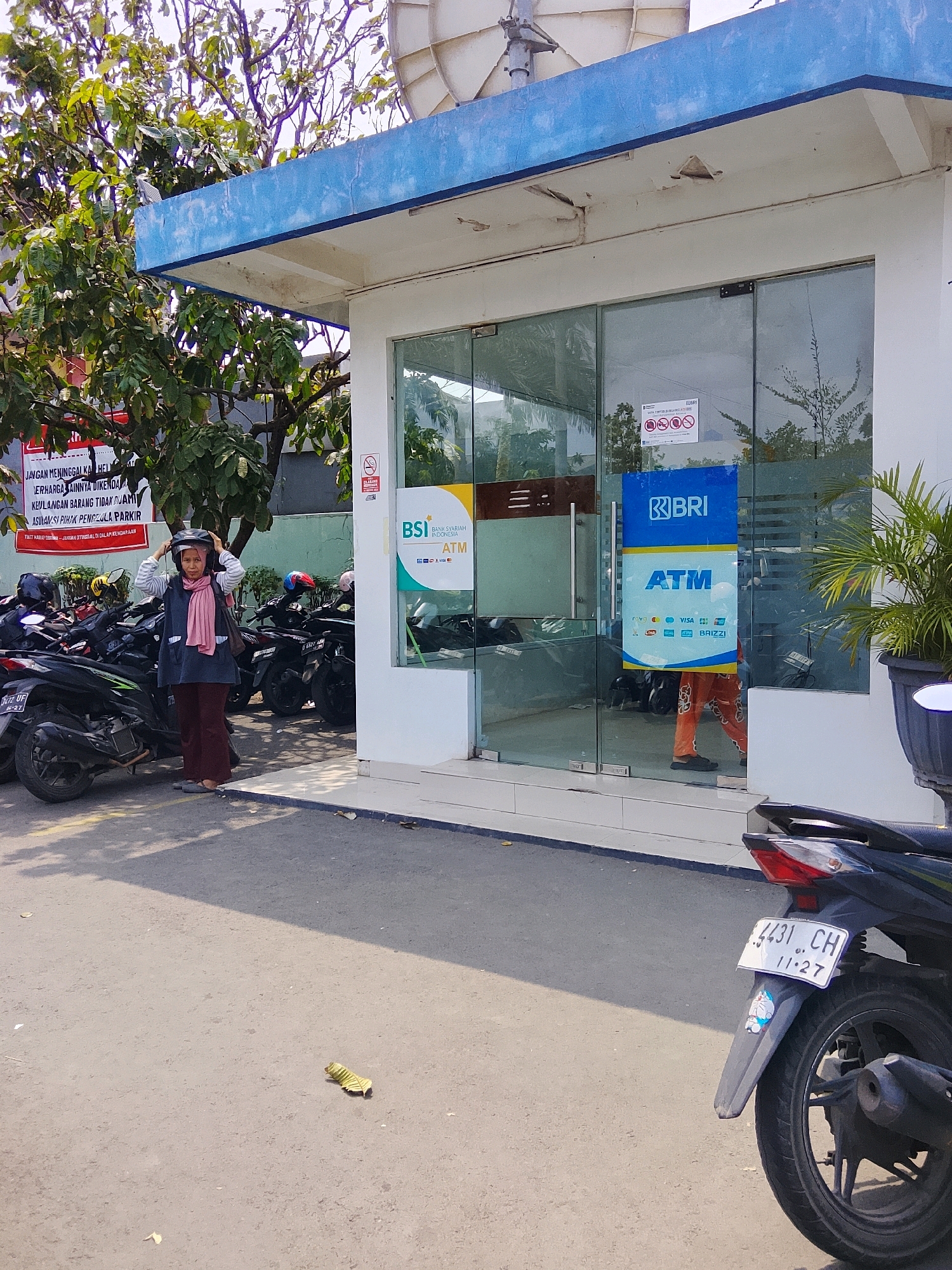 A woman wearing a pink scarf and a helmet is standing outside an ATM in Cirebon, Indonesia. She is facing the camera, with her left hand holding the helmet on top of her head. In front of her are several motorcycles parked in rows. The ATM is housed in a white building with a blue awning. The glass doors of the ATM have logos for BSI Bank Syariah Indonesia and BRI. To the right of the building, a large potted plant is visible. The ground is paved with asphalt, and a dry leaf is lying on the ground.