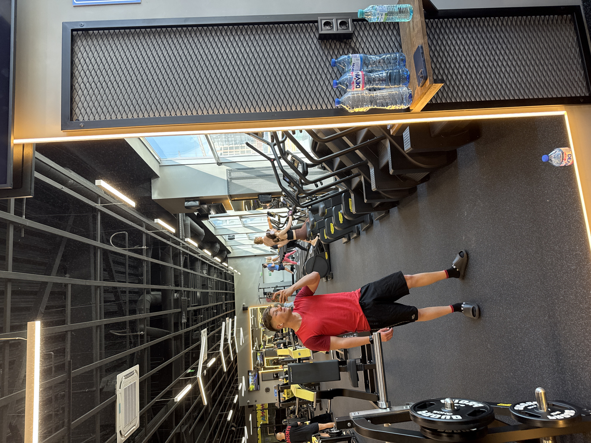 Here's a description of the image:
**Scene:** Interior of a gym, likely in Burgas, Bulgaria, inferred from the "DEVIN" water bottles visible in the photo. The gym appears to be modern and well-equipped.
**People:**
* A young man, dressed in a red t-shirt and black shorts, is taking a selfie. He is holding his phone and looking at the screen.
* Several other individuals are present in the background, using various gym equipment, including cardio machines (stair steppers).
* One person is on the bench and someone is walking by.
**Objects:**
* Various gym equipment, including weightlifting machines and cardio equipment (stair steppers).
* Weight plates on a weightlifting machine.
* Bottled water, with "DEVIN" labels, suggesting a Bulgarian location. Some bottles are on a shelf, and one is on the floor.
* The ceiling contains air conditioning components.
* The reflection of the buildings through the window shows the outside scenery.
**Actions:**
* The young man is taking a selfie.
* Other people are exercising on the gym equipment.
**Environment:**
* A modern gym with a high ceiling and ample lighting.
* The walls appear to be a mix of mirrored surfaces and decorative panels.
* The floor is dark.
* A window shows an outside view.