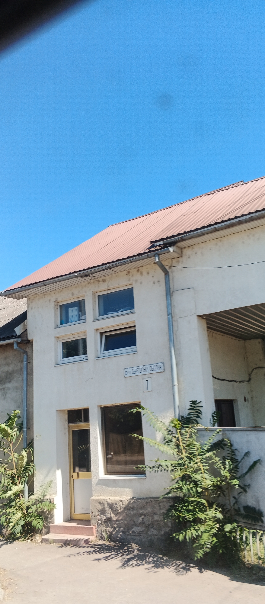 This is a photograph taken from a slightly elevated perspective, capturing the facade of a two-story building under a clear blue sky. The building has a light-colored, somewhat weathered exterior and a corrugated, reddish-brown metal roof. There are several windows visible on the upper floor, arranged in a grid of four. The lower floor features a doorway with a yellow-tinted glass door and a stone-clad foundation. A street sign with text and the number "7" is mounted on the wall. The building is surrounded by some greenery, with a tree or large shrub in the foreground and more foliage to the left and right. The ground in front of the building appears to be a paved surface.
This scene is located at vul. Berehova Ob'izna, 7 in the city of Mukachevo, Ukraine.