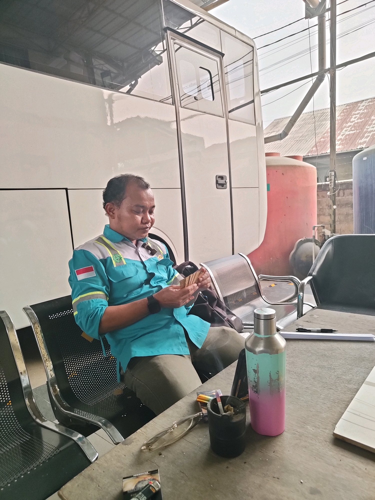 A man in a teal uniform with reflective stripes sits on a metal bench, holding what appear to be several bills in his hands. He is looking down at the money, his eyes partially closed. He wears a dark wristwatch on his left wrist. The man is seated in front of a large white vehicle, possibly a bus or a trailer. Behind the vehicle are large, red and blue cylindrical tanks. The environment appears to be an outdoor area with a concrete floor and some overhead structures. In the foreground, a wooden table holds a notebook, a pen, a water bottle with a gradient of teal and pink, an ashtray with a lit cigarette, and a pack of cigarettes. The man's uniform has an Indonesian flag patch on the left sleeve. The scene is set in an industrial or transport-related area, likely in the city of Maburai, Indonesia, given the presence of the Indonesian flag.