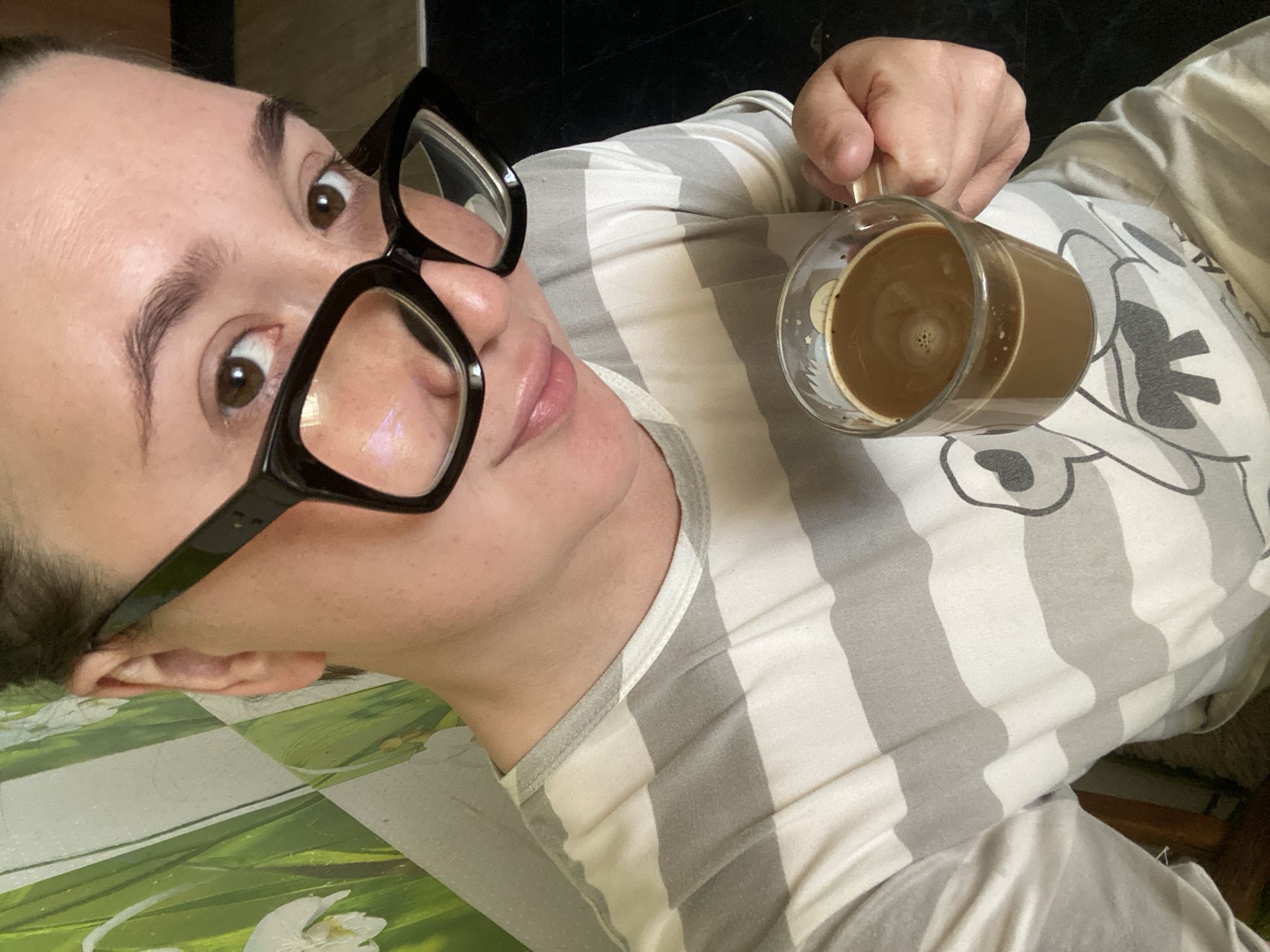 The image is a close-up, selfie-style shot of a person holding a glass mug of coffee. The person is wearing large, black-rimmed glasses and a gray and white striped t-shirt with a cartoon graphic on it. Their hair is pulled back, and their skin tone appears fair. The background shows a green and white patterned wall and a dark floor. The coffee in the mug is brown and has a foamy surface. No specific location in Chaikovsky, Russia can be identified from the visual information provided.
