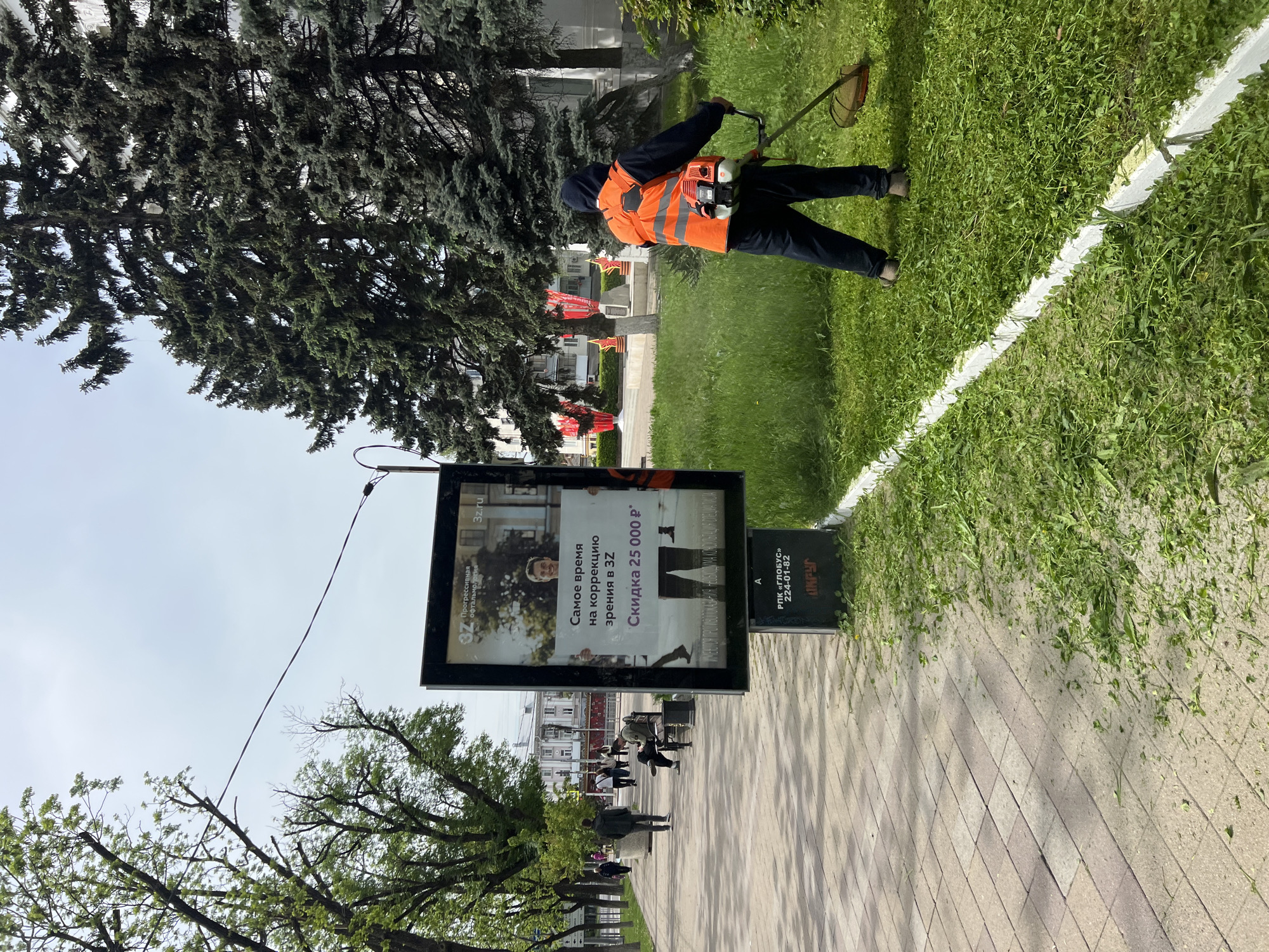 The image shows a street scene in Krasnodar, Russia. A man wearing a high-visibility orange vest is cutting grass with a string trimmer. The trimmed grass is scattered along a white painted line. An advertising board for an ophthalmology clinic, "3Z", promoting a vision correction discount is prominently displayed. Several pedestrians are visible in the background. The environment includes trees, a paved sidewalk, and buildings.