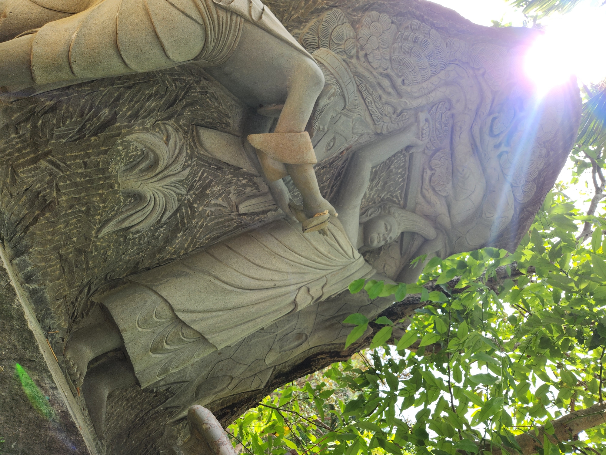 This image features a detailed bas-relief sculpture carved into a stone structure, likely a temple or monument, located in Đà Nẵng, Vietnam. The sculpture depicts two figures, one in the foreground on the left and another to the right. The figure on the left is kneeling or in a low crouch, with its left arm extended and hand grasping something held by the figure on the right. This figure wears a garment with layered details and a headdress. The figure on the right stands taller, draped in flowing robes, and is holding an object or supporting the other figure's gesture. Both figures appear to be engaged in a shared action or narrative. The sculpture is textured with various carved patterns, including foliage and intricate designs. Behind and above the sculpture, lush green foliage from trees creates a natural backdrop, with sunlight filtering through the leaves, casting bright rays and lens flares across the upper portion of the image. The stone has a weathered, aged appearance. The overall scene suggests a cultural or religious artwork set within a natural, outdoor environment.