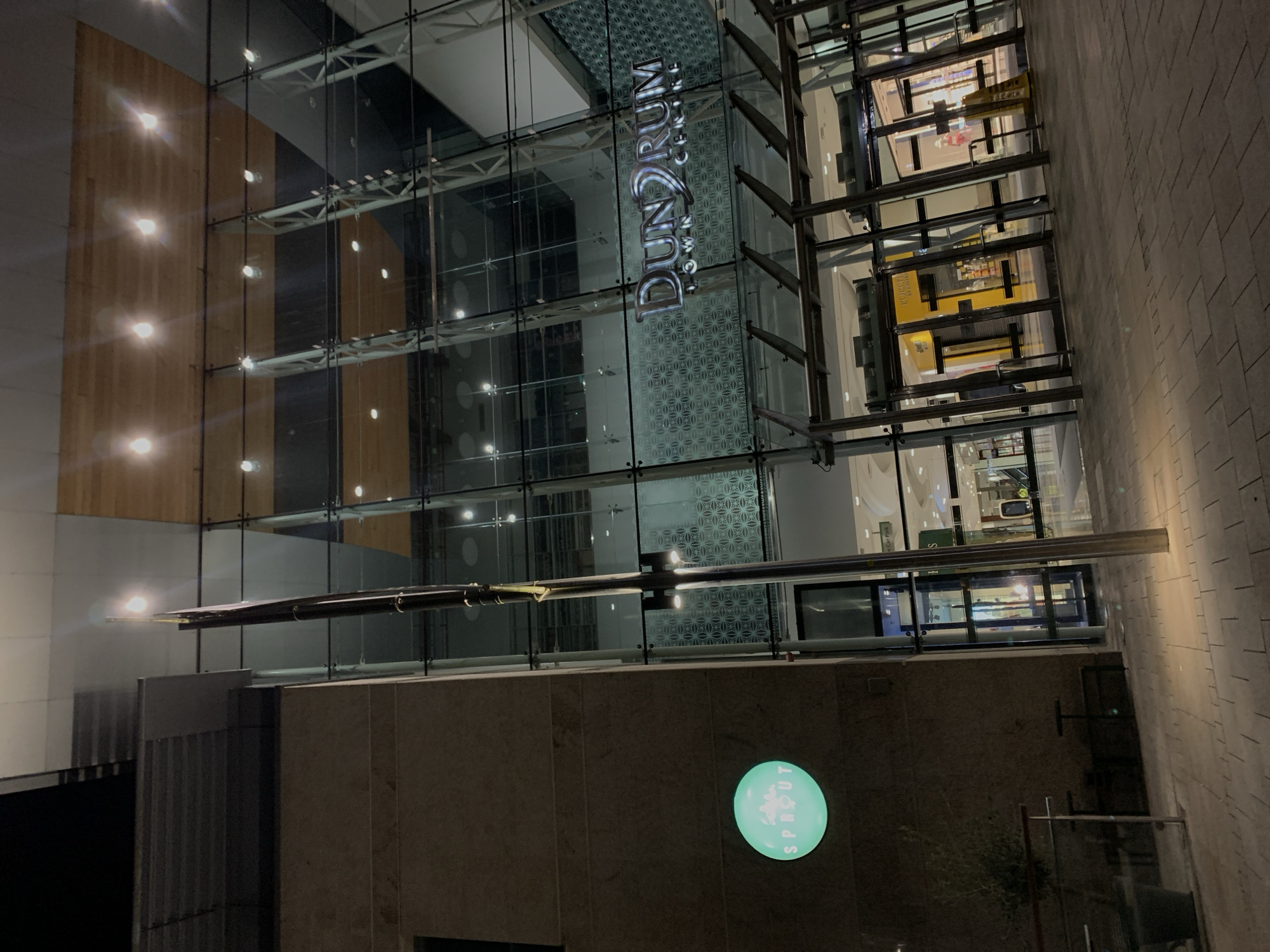 This is an exterior shot of Dundrum Town Centre in Dublin 16, Ireland, at night. The photo shows a multi-story structure with a glass facade displaying the "Dundrum Town Centre" sign. Below the glass facade is a stone-like wall with a circular, illuminated "Sprout" sign. The lighting appears dim, emphasizing the architectural lines and the signs. The exterior setting features a walkway paved with rectangular stone tiles. No people are visible in the scene.