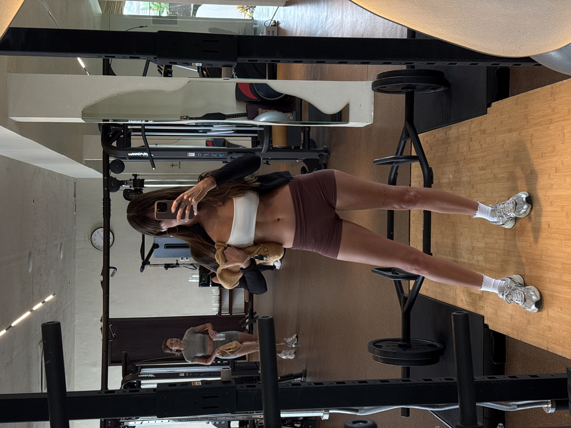 The image depicts a woman in athletic wear taking a selfie in a gym. She is wearing a white sports bra, a black zip-up jacket, and maroon shorts, along with white socks and silver athletic shoes. She holds a beige stuffed toy in her left arm. In the background, another woman in a grey athletic outfit is visible, holding a towel and a water bottle. The gym features weightlifting equipment, including a squat rack and barbells, and is illuminated by modern lighting fixtures. The overall environment suggests a well-equipped fitness facility. The location is identified as a gym in North Kuta, Indonesia.