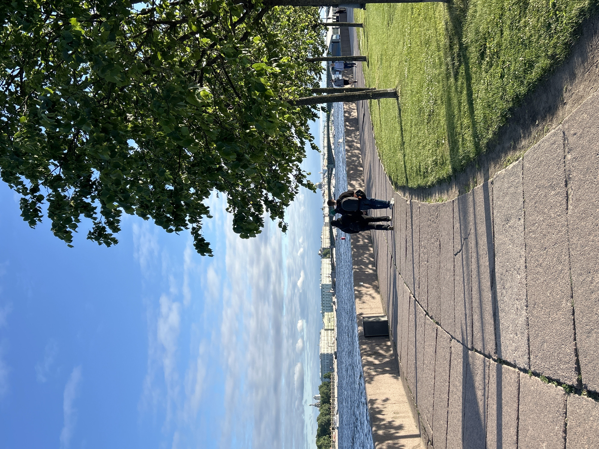 In this outdoor, daytime shot, a paved walkway curves along a body of water, with a grassy area to the right bordered by trees. Two individuals, facing away from the camera and walking towards the distance, are positioned on the walkway in the lower left quadrant of the image. The walkway is made of large, rectangular stone slabs with some growth visible in the cracks. The water is choppy, and in the background, a long, low building complex with a light-colored facade stretches along the opposite bank. A bridge is visible in the distance, and a few boats are on the water. The sky is bright blue with scattered white clouds. The trees casting shadows on the walkway are lush and green.