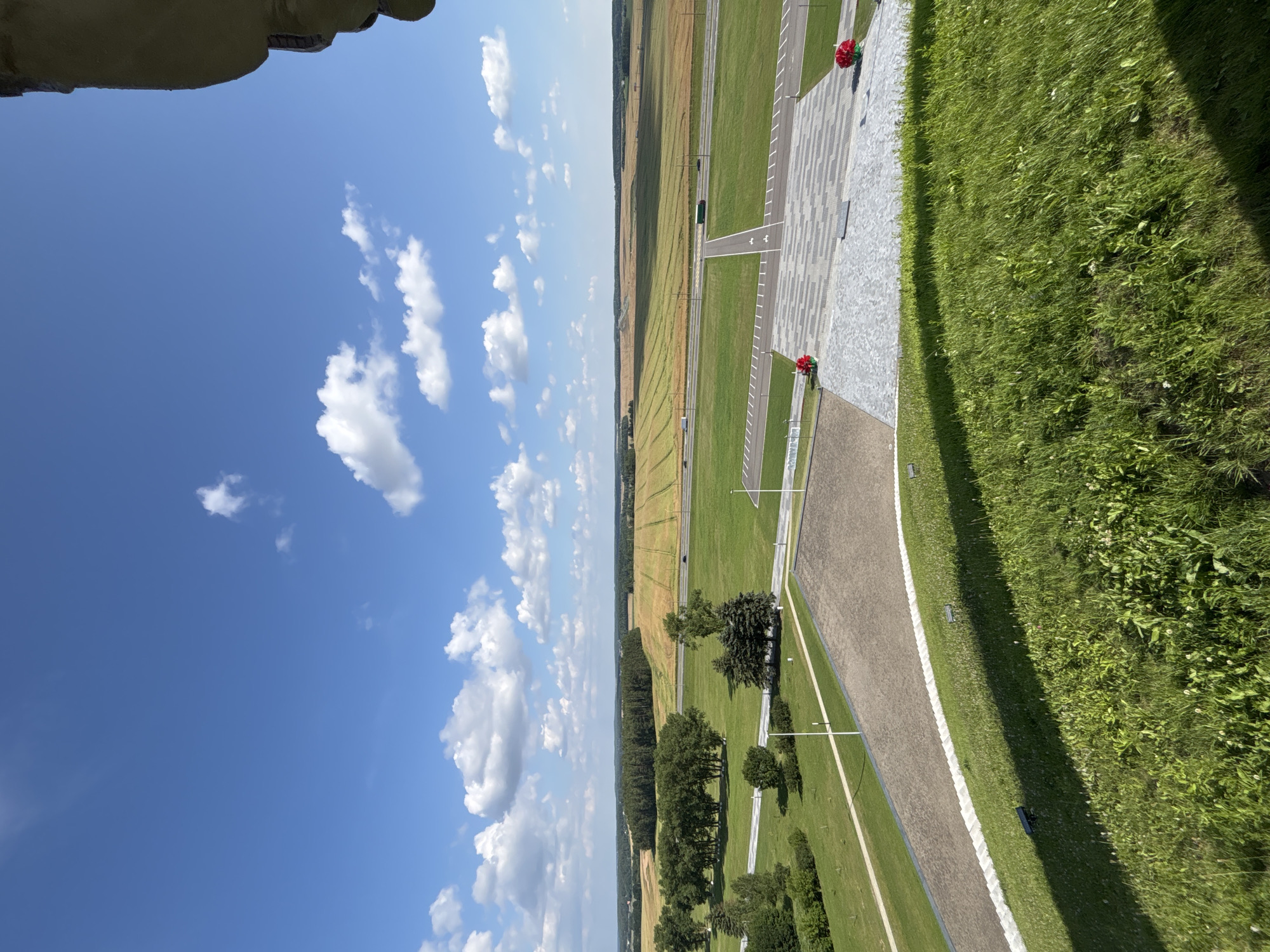 A high-angle, wide shot captures a sunlit landscape under a bright blue sky with scattered white clouds, likely in the Ozyaritska-Slabadski rural council, Belarus. The foreground is dominated by lush green grass, casting a shadow from an unseen object. Beyond the grass, a paved area with a patterned surface leads to a path. Further in the mid-ground, a mowed green field stretches towards a highway with two vehicles, one a white truck and the other a green truck, and a line of trees bordering the field. In the background, rolling hills are covered in a golden-yellow crop, suggesting a rural setting. To the right edge of the frame, a portion of a large, textured, beige-colored structure, possibly a monument or building, is visible.
