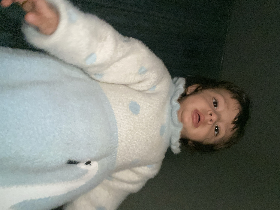 A baby lies on a dark surface, likely a couch or bed. They are wearing a fuzzy, light blue and white romper with blue polka dots and a ruffled collar. The baby's right arm is extended upwards, and their mouth is slightly open. The infant has dark hair and fair skin. The lighting is dim, casting shadows across the background.