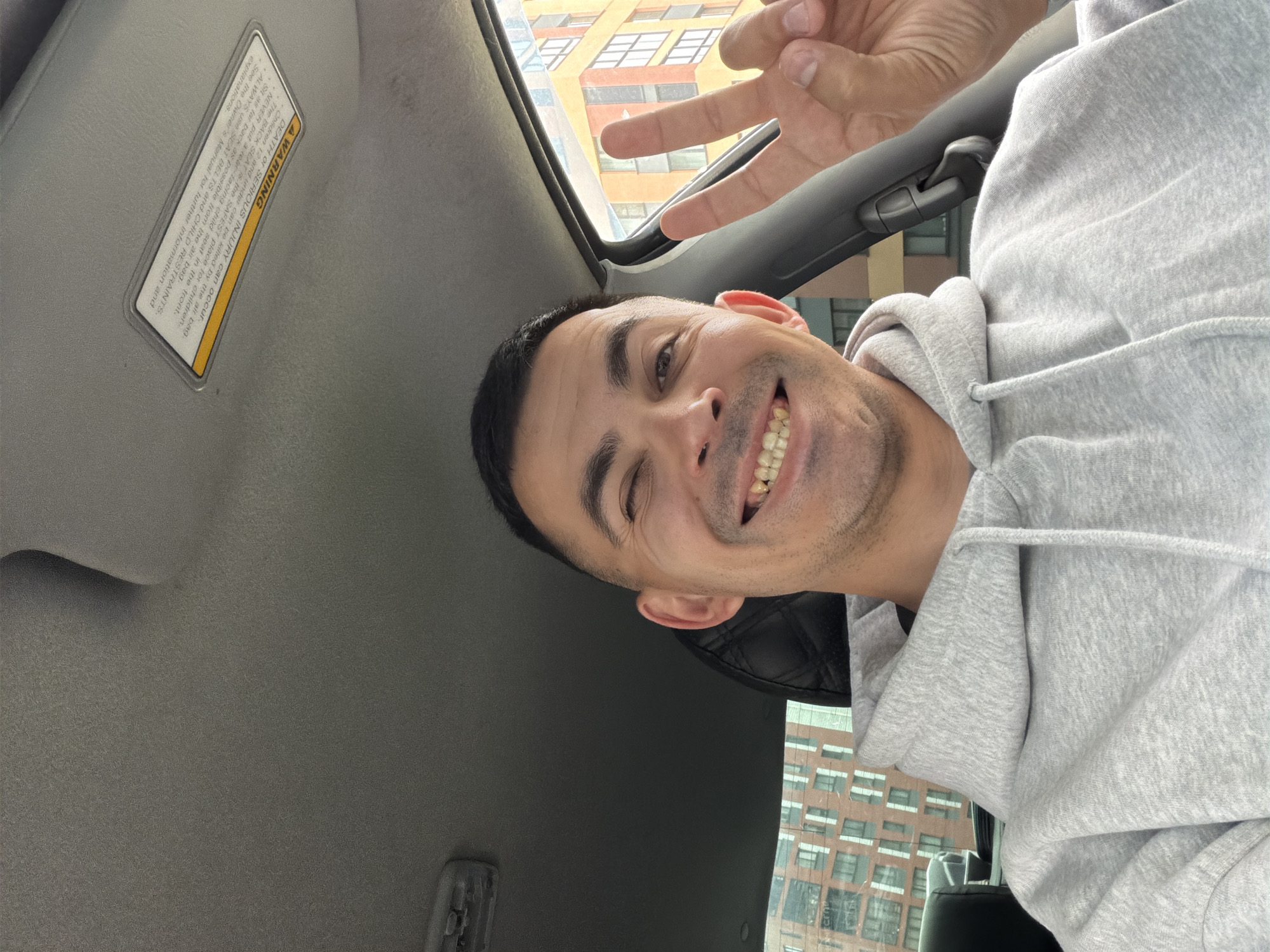 A man takes a cheerful selfie inside a car, likely in Izhevsk, Russia. He is positioned on the right side of the frame, smiling broadly with his teeth visible, and appears to be winking or raising an eyebrow. He wears a light gray hoodie, and his right hand is raised in the upper right corner, making a peace sign.
The setting shows the interior of the car, with a light gray headliner above him and a sun visor in the upper left. Through the car window or sunroof, a multi-story building with numerous windows is visible, indicating an urban environment bathed in daylight.
A detailed warning label on the sun visor is clearly legible. The text reads: "A WARNING. DEATH or SERIOUS INJURY can occur. The BACK SEAT is the SAFEST place for children. Children 12 and under can be killed by the air bag. NEVER put a rear-facing child seat in the front. ALWAYS use SEAT BELTS and CHILD RESTRAINTS. Sit as far back as possible from the air bag. See the Owner's Manual for further information and explanations."
The man's happy expression and casual gesture suggest a lighthearted moment captured in a self-portrait.