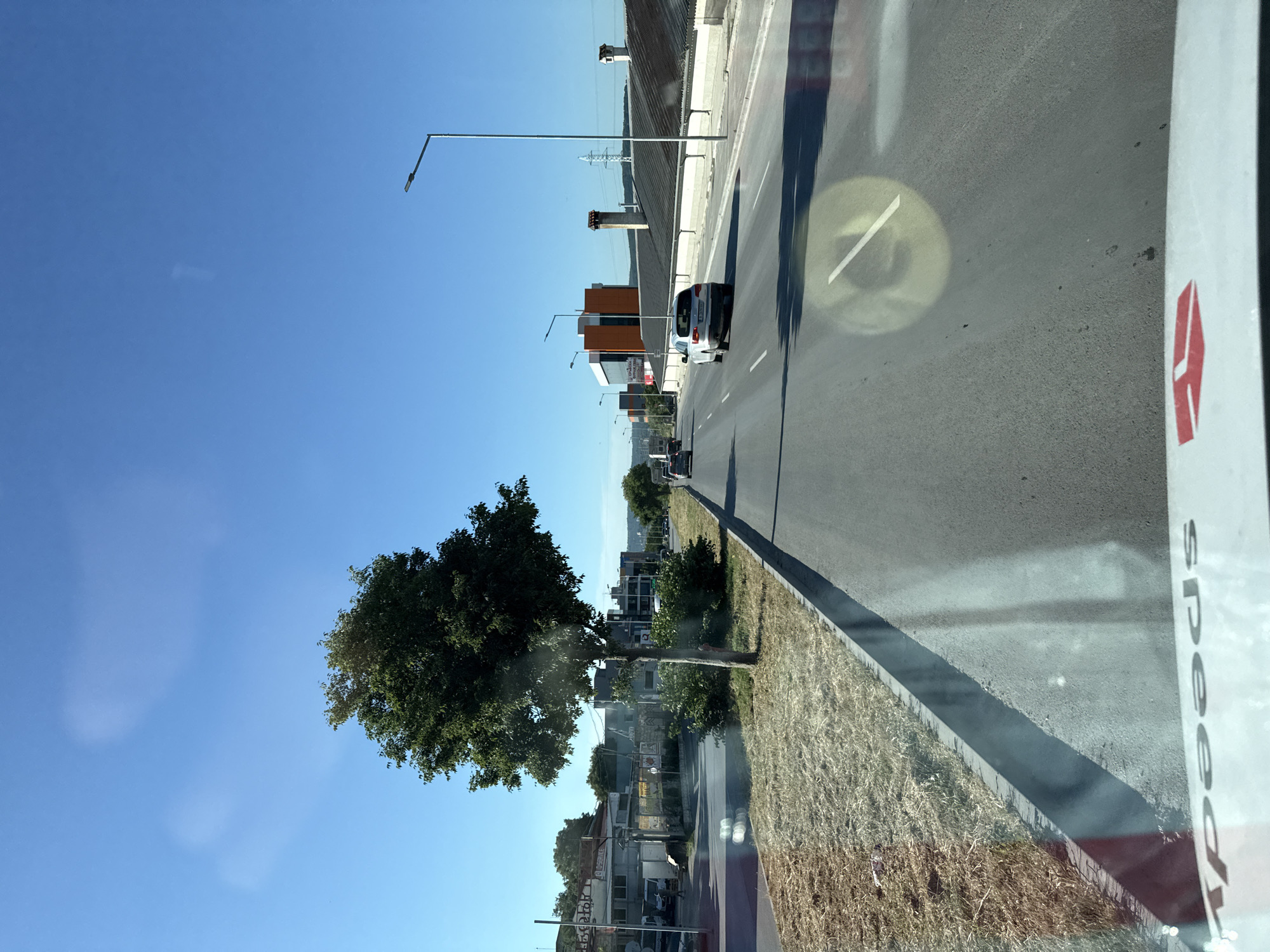 The image shows a view from a vehicle traveling on a multi-lane road in Varna, Bulgaria, likely on a sunny day given the bright blue sky. On the left side of the road, there is a dry, grassy embankment and a lone tree with full green foliage. Further left, buildings are visible, some appearing to be commercial or industrial, with signage in a language that appears to be Korean and English. Vehicles are driving on the road, including a white SUV in the right lane. On the right side of the road, there are buildings with dark, sloping roofs, and chimneys. Streetlights are visible along the road. The road surface is asphalt, and there are lane markings. The foreground of the image shows a portion of the vehicle's dashboard, with the word "Speedy" printed on it, indicating it is likely a courier or delivery vehicle. There are reflections on the windshield of the vehicle. The overall environment suggests an urban or semi-urban area, possibly on the outskirts of Varna, with ongoing traffic and commercial activity.