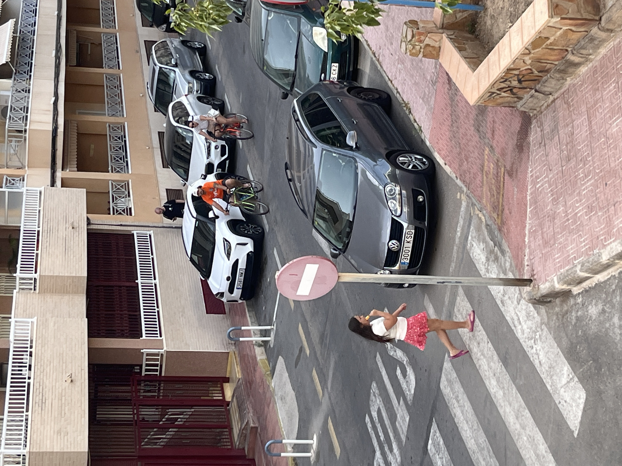 The image depicts a street scene in Torrevieja, Spain, likely on a residential street. In the foreground, a young girl wearing a white t-shirt and a pink skirt is walking across a pedestrian crossing, holding what appears to be a piece of food. She is wearing pink flip-flops. A traffic sign with a red circle and a white bar stands nearby. Several cars are parked on the street. A dark grey Volkswagen Golf station wagon is prominently featured in the foreground. Behind it, a green hatchback is parked. Further down the street, a silver SUV and a white SUV are visible, with two individuals on bicycles in front of them. One cyclist wears an orange shirt, and the other is wearing a striped shirt. A man is standing near the white SUV. The buildings in the background are typical of Spanish coastal architecture, with balconies and beige brick facades. The street is paved, with white lines marking the pedestrian crossing and yellow lines indicating traffic lanes.