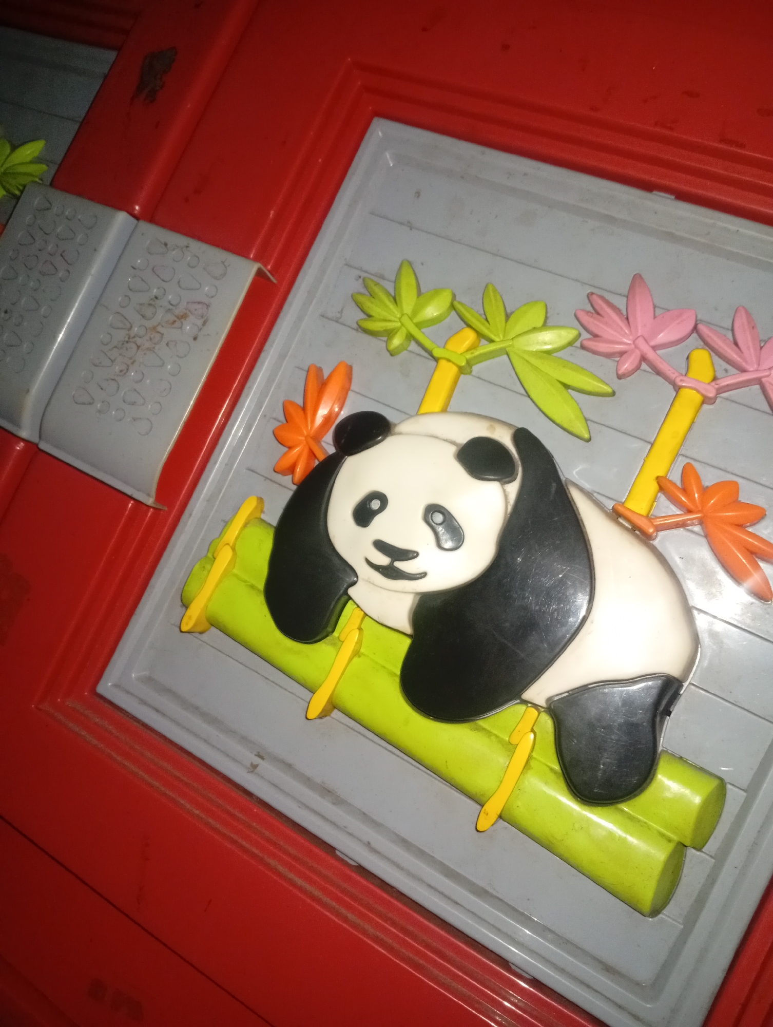 A close-up shot captures a plastic toy panda perched on bamboo stalks against a gray background. The panda is white and black with a smiling expression, and it's surrounded by colorful plastic leaves in green, pink, and orange. The toy is set within a red frame. The image shows signs of wear and tear, with scuffs and dust visible on the toy and its surroundings. The scene is dimly lit, suggesting it might be an indoor setting. The precise location of this scene in Nyalindung, Indonesia, cannot be determined from the provided image alone.