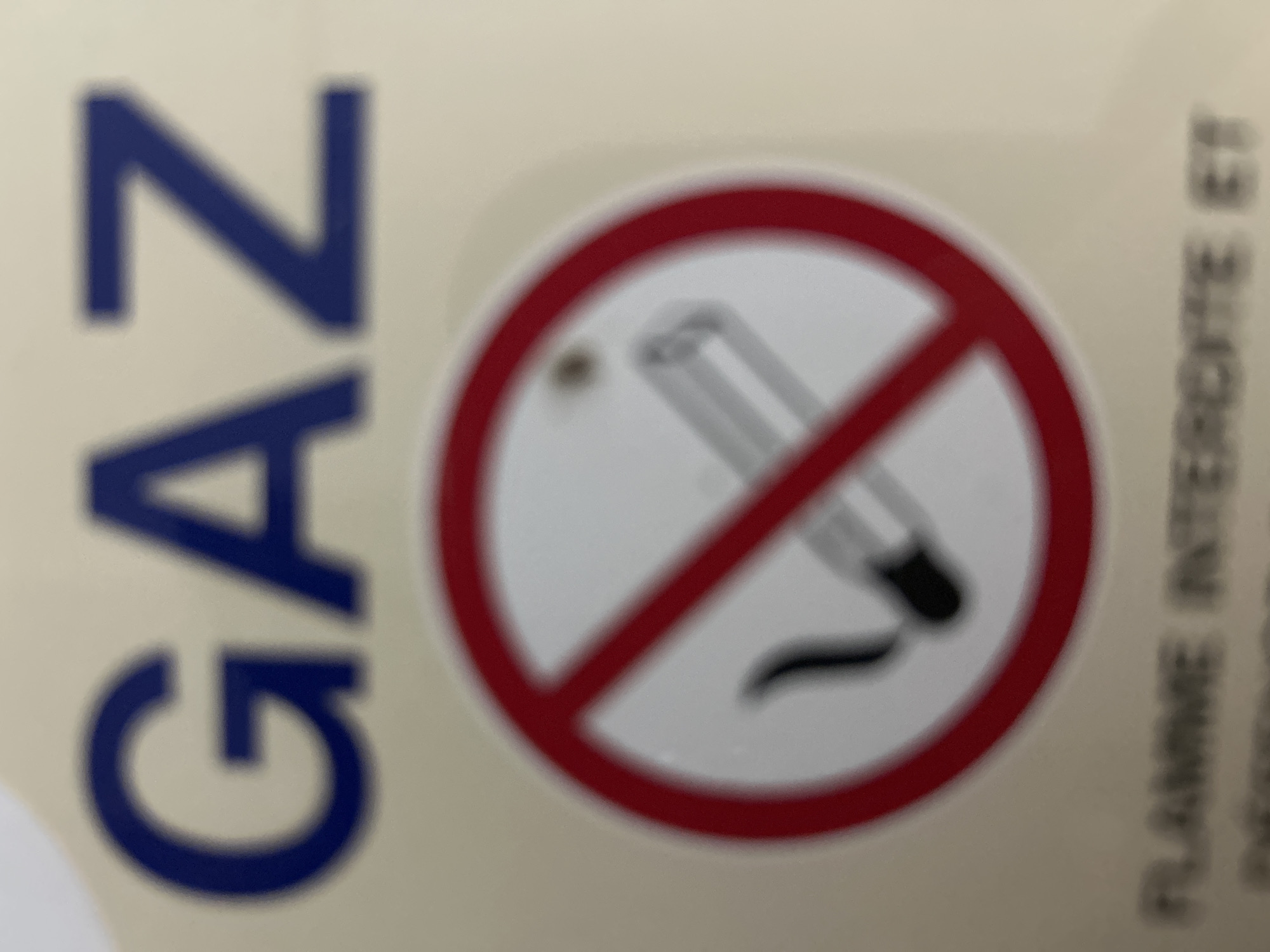 The image shows a sign with the word "GAZ" in large blue letters at the top. Below "GAZ" is a circular red prohibition symbol with a diagonal line through it. Inside the circle is an illustration of a cigarette with smoke, indicating no smoking. At the bottom of the image, text is partially visible, reading "FLAMME INTERDICTE ET". The background is a light yellow or beige. The sign appears to be located in a public area, possibly a building or facility where gas is present and smoking is prohibited. No specific location in Anduze, France is discernible from the image itself.