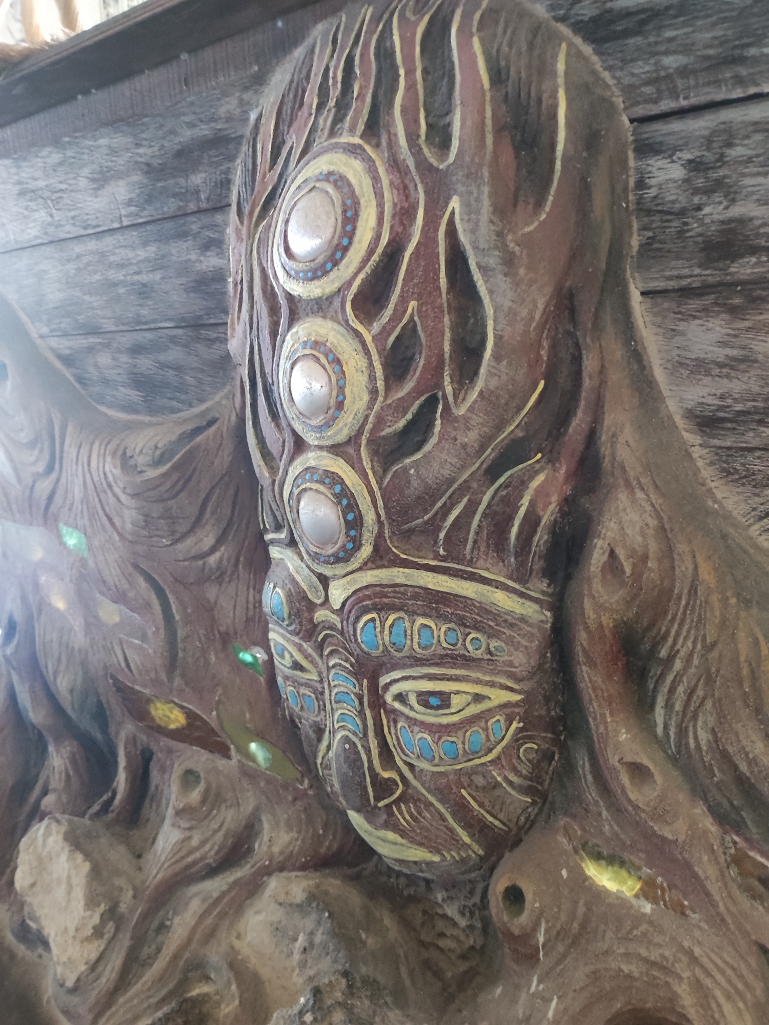 A close-up shot captures a carved wooden mask with an intricate design. The mask, predominantly brown, features three large circular elements along its vertical axis, each surrounded by a yellow ring dotted with blue. Below these, the mask's face exhibits more detailed carving, with eye-like shapes decorated in yellow and blue, and stylized lines forming other facial features. The wood around the mask appears textured and rough, adding to its rustic, handcrafted appearance. The background includes weathered wooden planks. The location is Unknown, Indonesia.