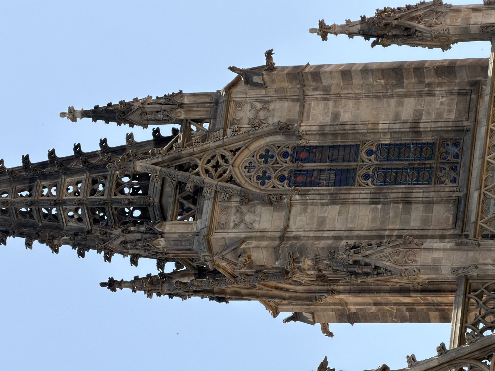 This is a close-up, eye-level, outdoor photograph of the upper section of the Cathedral of Barcelona, Spain. The focus is on the intricate Gothic architecture of the bell tower and its spires. The stone structure is weathered and textured, with elaborate carvings, gargoyles, and pointed arches. A large stained-glass window, featuring geometric patterns in shades of blue and red, is visible on the tower. Several smaller spires and decorative elements surround the main tower, all against a clear blue sky. There are no people or animals visible in the frame, and no discernible actions are taking place. The lighting suggests a sunny day.
