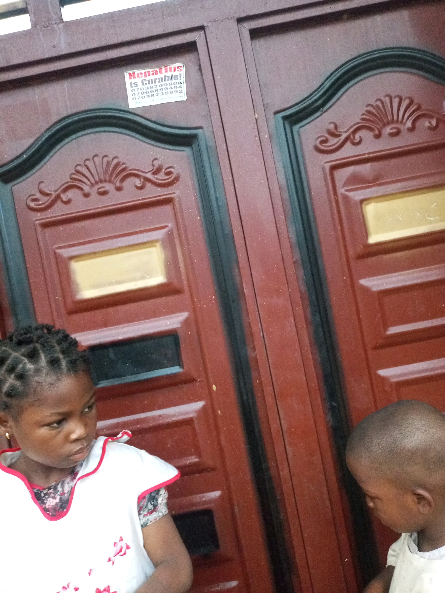 A medium shot captures two young Black children standing in front of a maroon metal door in Tempure, Nigeria. On the upper left of the door, a sticker reads "Hepatitis Is Curable!" followed by three phone numbers. The girl on the left wears a white top with pink trim and a patterned sleeve, her hair in cornrows. The boy on the right, with a shaved head, is partially visible on the right side of the frame. The door features raised paneling and decorative elements.