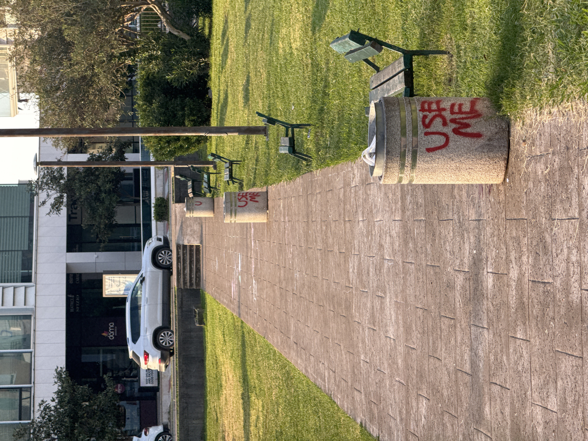 This outdoor image captures a paved pedestrian pathway flanked by green lawns, suggesting a park or public area in Budva, Montenegro. The scene is illuminated by warm, low-angle sunlight, indicating either early morning or late afternoon, with distinct shadows cast across the grass.
Along the right side of the pathway, several concrete trash bins are visible, each with a metal band near the top. Two of these bins prominently display the words "USE ME" in red spray paint. Adjacent to the trash bins are green metal benches with wooden slats, facing away from the path. On the left side, tall poles run parallel to the pathway.
Beyond the lawns, various trees and dense green bushes add to the natural elements. In the background on the left, modern multi-story buildings with large windows and white facades are visible. A white SUV is parked near one of these buildings, and a set of concrete stairs leads upwards behind it.
Visible text on the buildings includes "doma" (appearing twice), "BEATRICE.", "SFIZIO", "elena", and "CARAC". A sign near the parked SUV partially reads "KEEP SMILING AND ENJO GOOD FOO", likely implying "ENJOY GOOD FOOD". There is also a partial word "Tra" visible. No specific activity is taking place, presenting a quiet, urban landscape.