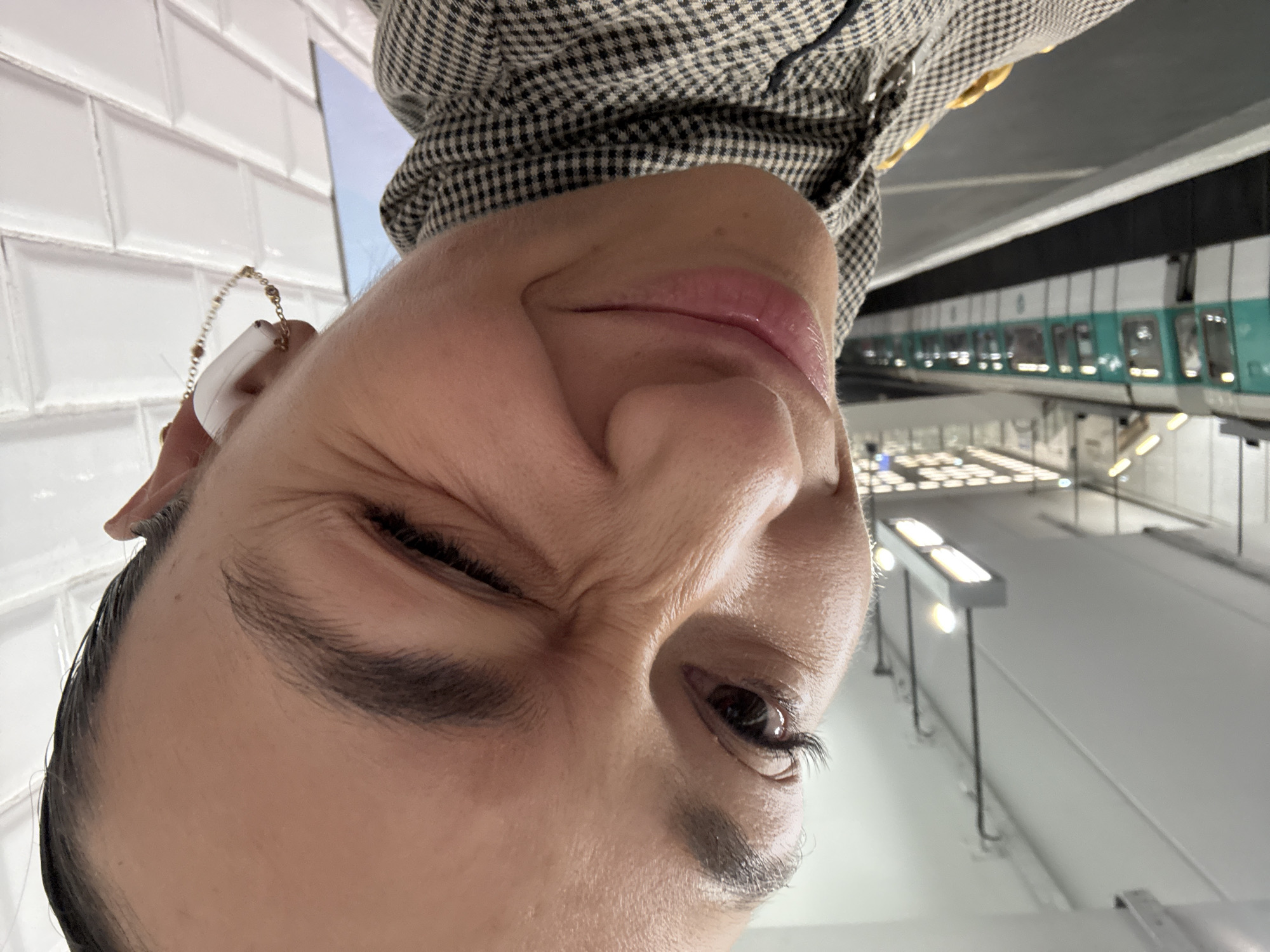 A person wearing a houndstooth patterned shirt is taking a selfie inside a Clichy, France metro station. The person is smiling and winking. A train is visible in the background. White tiles line the wall. Overhead lighting fixtures are also visible. The person has an earpiece with a beaded chain hanging from it.