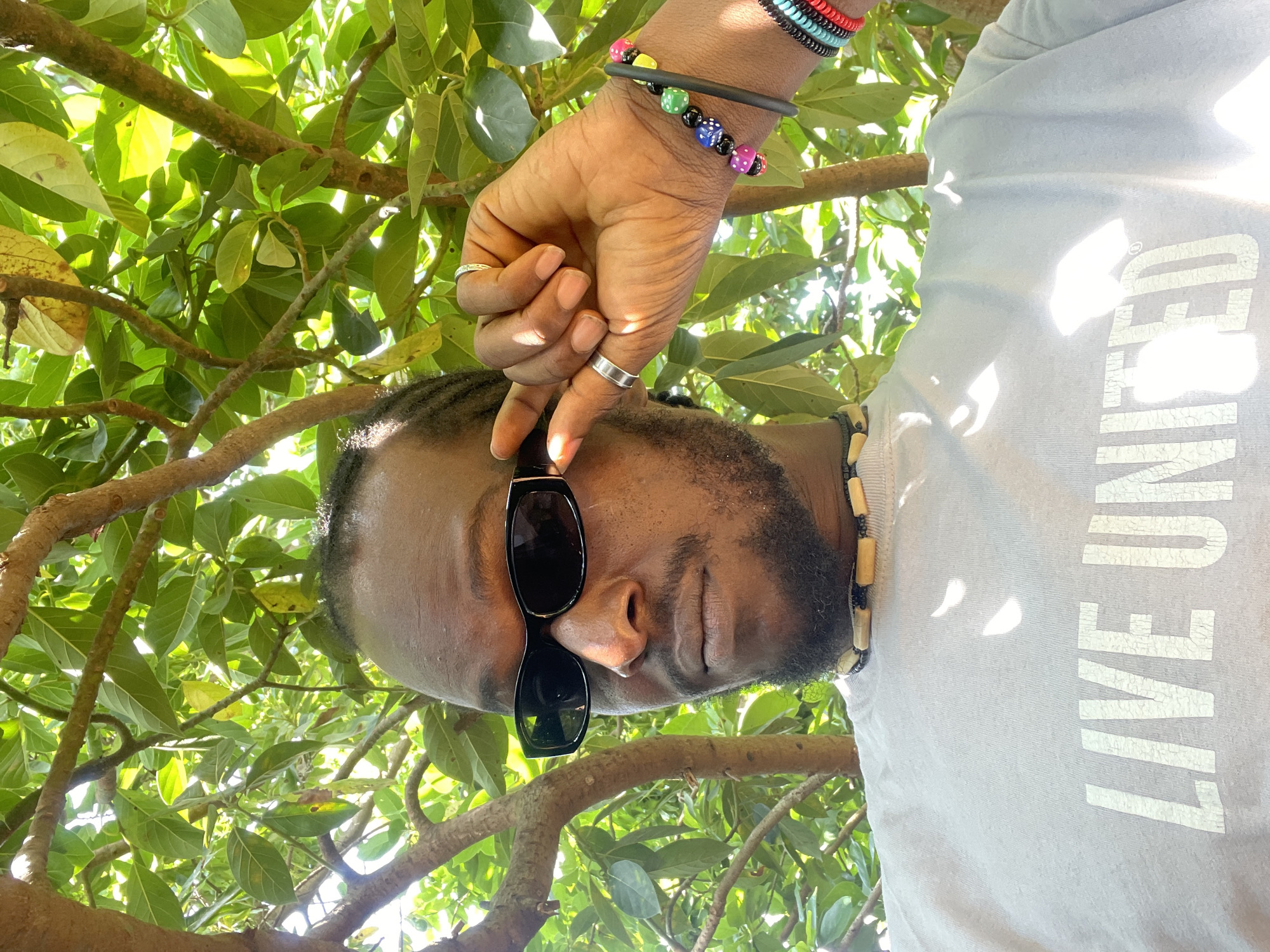 A man with dark skin, braided hair, a beard, and sunglasses is looking to his left. He is wearing a light grey t-shirt with the words "LIVE UNITED" printed on it in white. Around his neck is a beaded necklace, and on his right wrist, he wears several colorful bracelets. He is holding the frame of his sunglasses with his right hand, which also has two rings on it. The man is outdoors, with lush green leaves and tree branches visible in the background, suggesting a sunny day. The overall impression is a casual, outdoor portrait. The image does not contain specific landmarks that would allow for a precise location within Jos, Nigeria, but the abundance of foliage and sunlight points to a warm, tropical or subtropical climate, consistent with Jos.