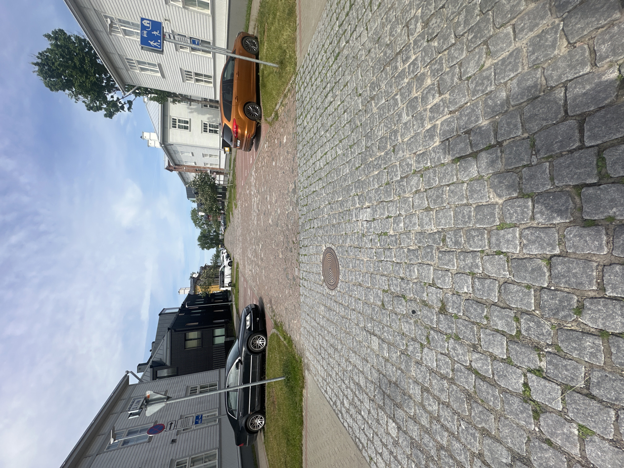 The image depicts a cobblestone street in Tallinn, Estonia, on a partly cloudy day. The perspective is from street level, looking down a gently sloping street lined with buildings on both sides.
On the left, a light gray wooden building with white trim and multiple windows occupies the foreground. A black sedan is parked on the grass verge in front of it. Further down the street on the left, a modern black building with dark windows stands next to the gray one.
On the right, a white wooden building with numerous windows stretches along the street. An orange SUV is parked in a designated parking spot in front of this building. Further down on the right, more white buildings are visible, along with trees and greenery.
The street itself is paved with grey cobblestones, with some grass growing between them. A circular metal manhole cover is visible in the foreground. Traffic signs are present on both sides of the street, including a blue square sign with a white silhouette of a person walking and a car.
The sky is a mix of blue and white clouds. The overall impression is that of a well-maintained urban environment, likely a residential or historical district within the city.