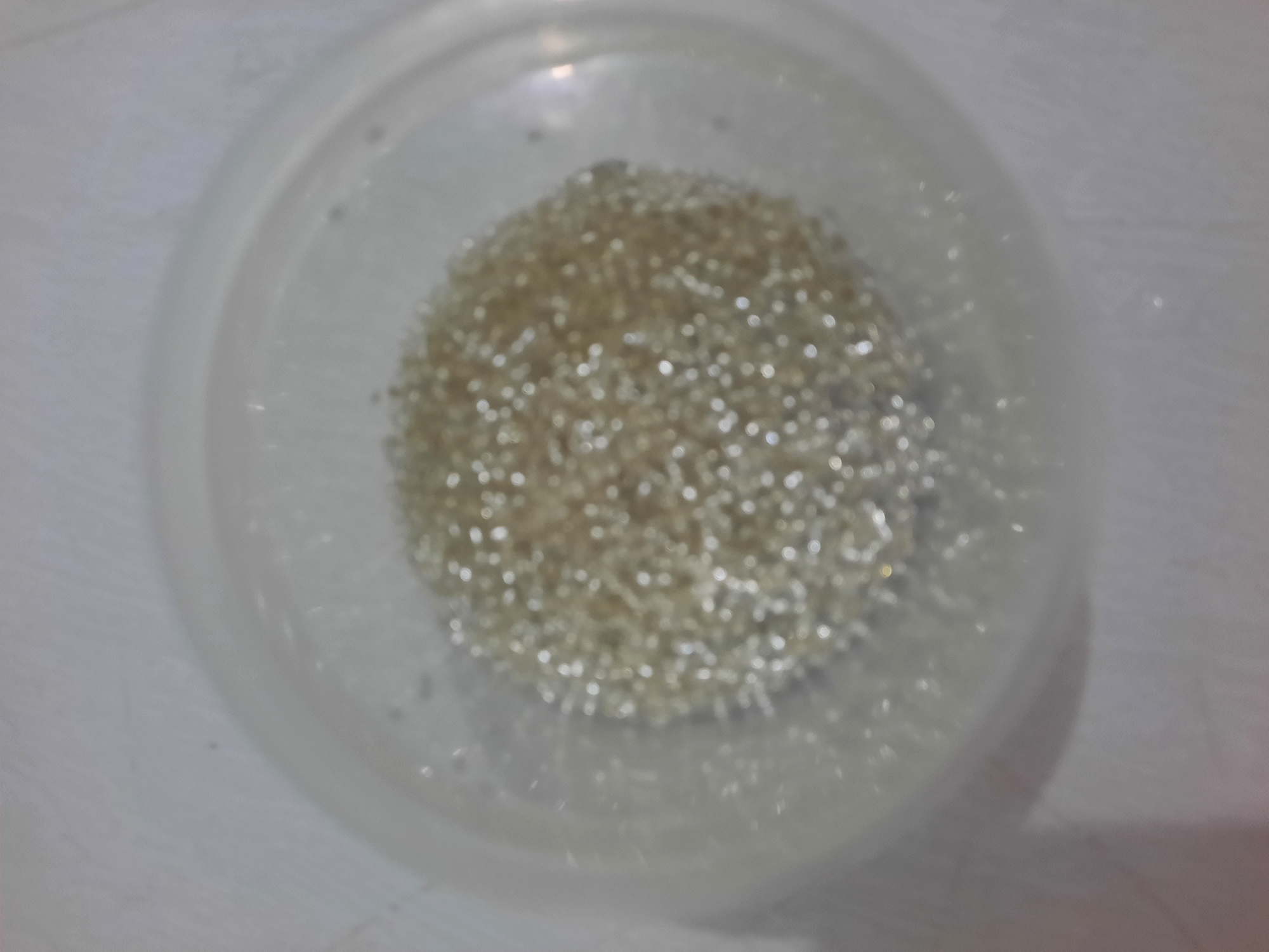 The image shows a close-up, overhead view of a clear plastic container filled with small, round, translucent beads. The beads are clustered in the center of the container and appear to be gold or clear in color, reflecting light. The container is round and has a slightly raised rim. The background is a light-colored, textured surface, possibly a table. There are no people or discernable landmarks visible in the image. The scene depicts a simple collection of beads in a container, with no specific context provided about its location in Lhokseumawe, Indonesia.