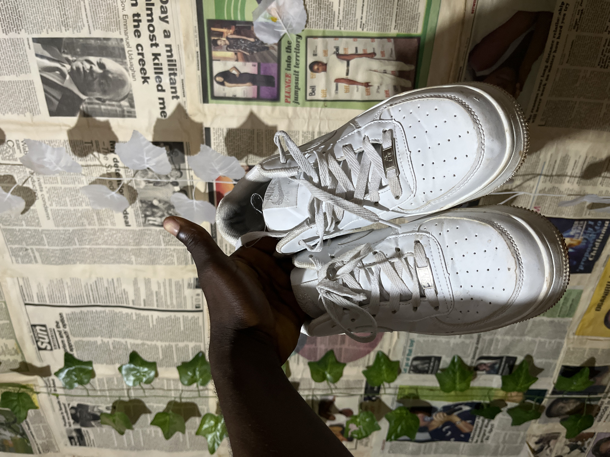 A pair of white Nike Air Force 1 sneakers is held up by a dark-skinned hand against a wall covered in what appears to be newspaper clippings and various printed materials. Artificial green ivy vines are attached to the wall, some casting shadows. The sneakers show signs of wear, with minor scuffs and dirt visible on the soles. The background newspaper articles are mostly out of focus, but some headlines and images are discernible, suggesting a newsstand or a decorated wall in an urban setting. The overall lighting is somewhat dim, with shadows contributing to the visual texture. No specific location within Rafin Par, Nigeria, is indicated by the visual evidence.