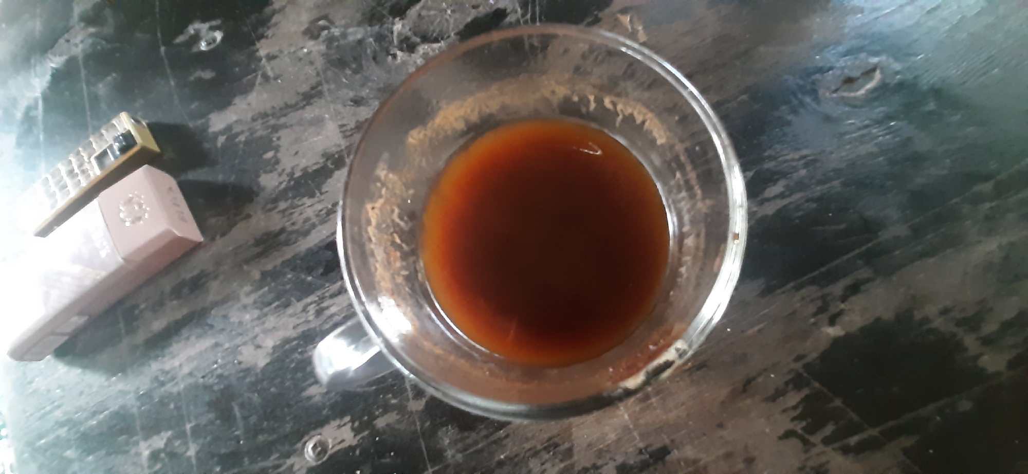 In Yessentukskaya, Russia, on a weathered, dark wooden table, a clear glass mug filled with a dark liquid, likely coffee or tea, sits centrally. To the upper left of the mug, a pack of cigarettes and a golden lighter are positioned. The table surface exhibits wear and tear, with visible grain and scuff marks. The lighting appears to be ambient, casting a subtle glow on the scene. No individuals are present in the image.