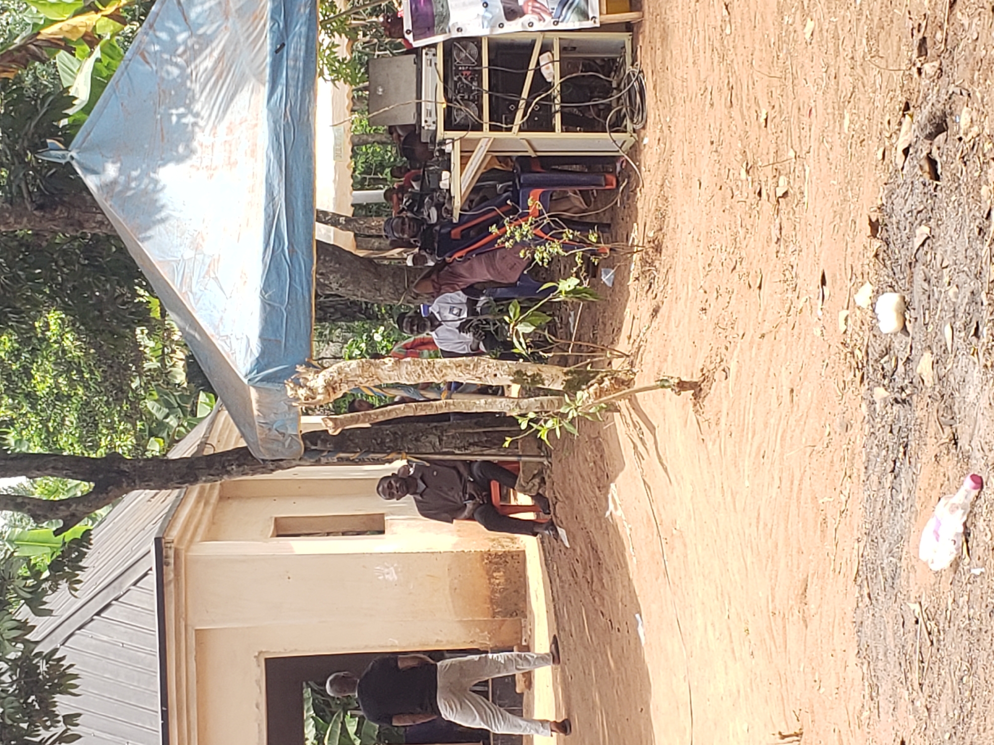 This image depicts an outdoor gathering in Umundugba, Nigeria, under bright daylight conditions. The scene is set on dusty, reddish-brown ground with ample green foliage and trees in the background.
On the right side of the frame, several individuals are seated on a mix of plastic chairs (orange and blue) beneath a blue tarp that serves as a makeshift shelter. Behind these seated people, a complex sound system or electronic setup is visible on a stand, featuring numerous wires and various components. A banner or poster, displaying what appears to be a portrait of a person, is affixed above the sound equipment.
In the mid-ground, a man dressed in a dark shirt and dark trousers sits on an orange chair, looking towards the viewer. To the left and slightly closer to the foreground, another man, wearing a dark shirt and light-colored trousers, stands with his back to the camera, near a light-colored building. This building has a dark window or doorway and a corrugated metal roof, partially obscured by tree branches.
The ground shows patches of varying texture and color, and a white plastic bottle with a pink cap is discernible among the debris in the foreground. Strong shadows cast by the sun indicate it is likely midday or early afternoon. The overall impression is of a community event or informal meeting in a rural or semi-urban environment. No explicit text is clearly legible in the image.