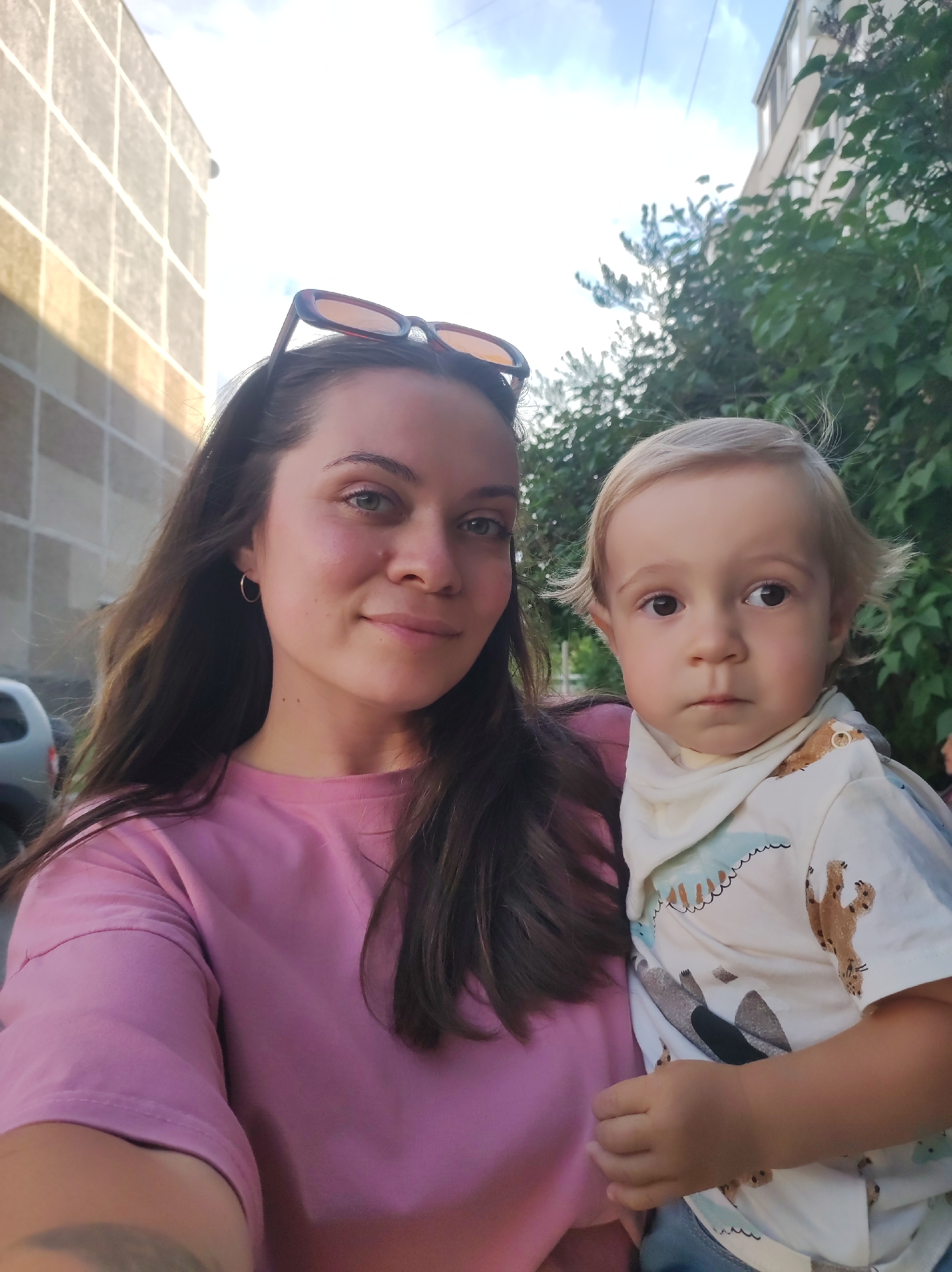 A woman and a young child are taking a selfie outdoors. The woman, with long dark hair and wearing a pink t-shirt, is on the left side of the frame, holding the child. She has sunglasses on top of her head and is smiling. The child, with blonde hair and wearing a light-colored shirt with animal prints and a bib, is held on the woman's right arm, facing forward. The child has a neutral expression.
The background shows an urban setting with buildings and trees. A building with a grid pattern is visible on the left, and a taller building and lush green foliage are on the right. The sky is visible above, with some clouds. The lighting suggests it is daytime, likely late afternoon or early evening, given the warm tones. There is no visible text in the image. The overall mood is casual and warm, capturing a moment between the woman and the child. The location context is Chaikovsky, Russia.