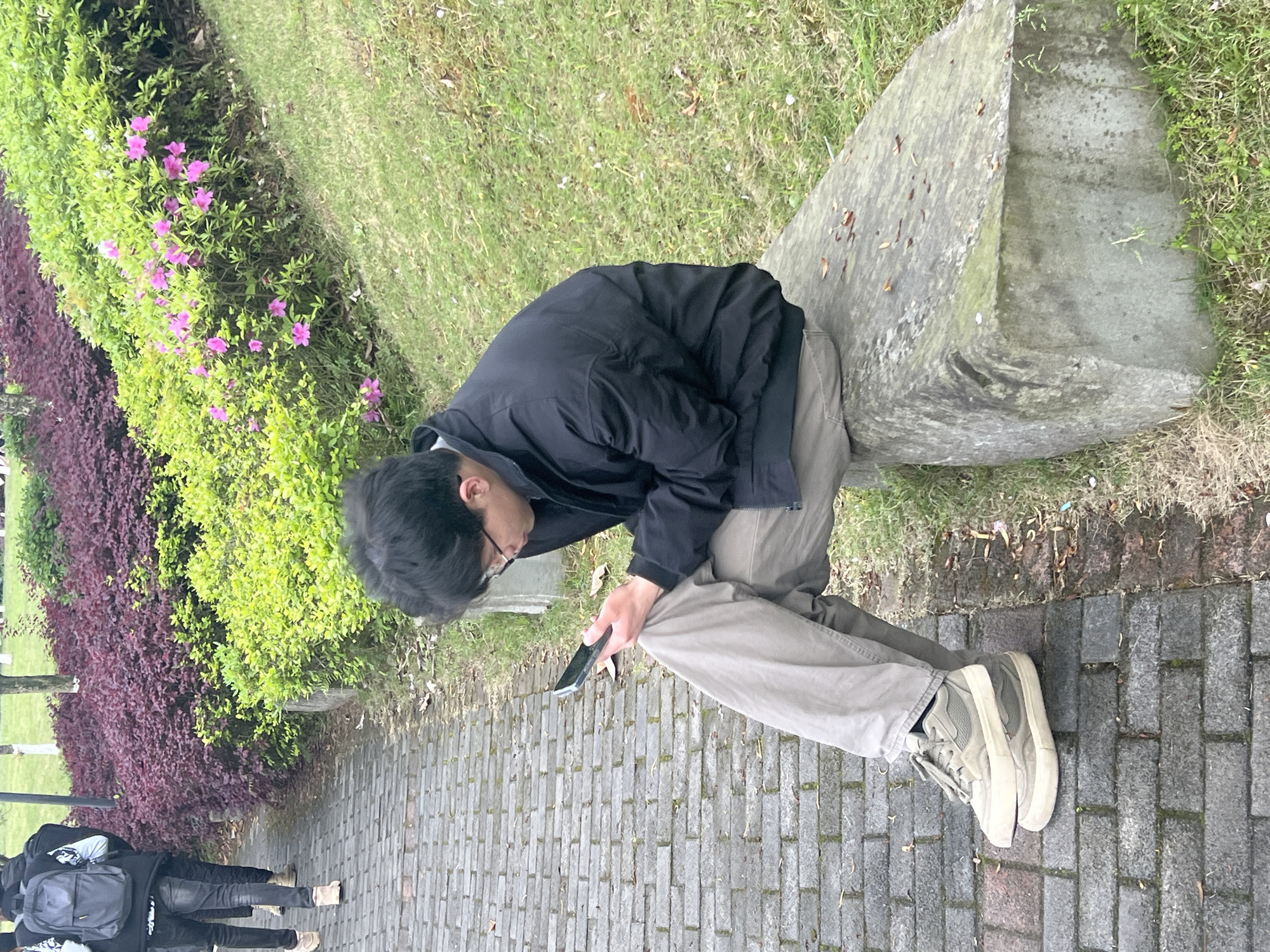 A young Asian man is sitting on a stone structure in a park. He is wearing glasses, a black jacket, and light tan pants, and is looking down at a smartphone in his hands. He is positioned on a stone-paved path with greenery to the left and grass surrounding the stone structure on which he sits. In the background, a group of people are walking along the path. Based on visual evidence, the location is Jiangjin, China.