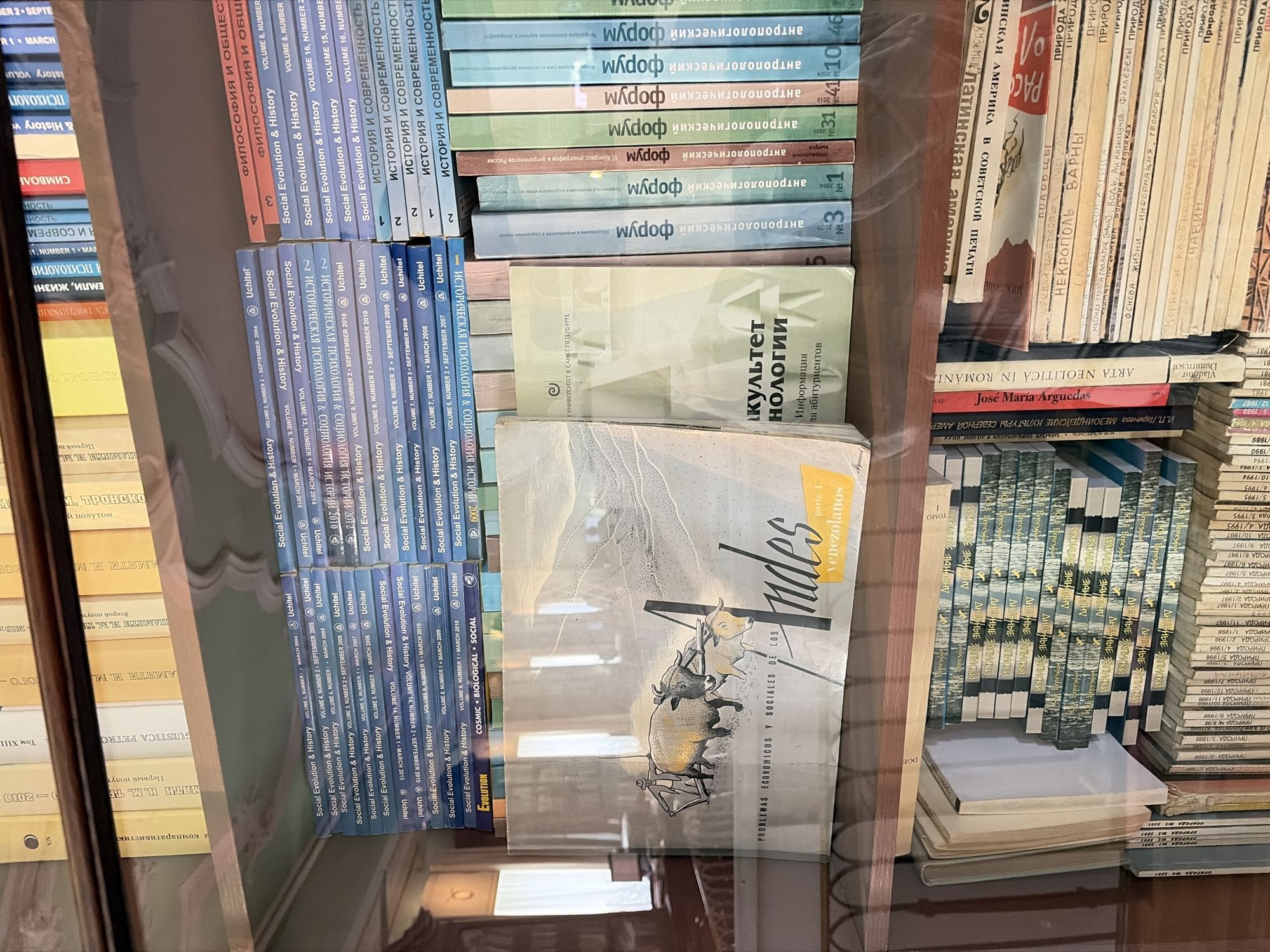 This is a photograph of several bookshelves filled with various books and journals. The volumes are densely packed, with titles in both Russian and English. Some of the English titles are "Social Evolution & History," and there are multiple volumes of "ДНЕВНЫЕ ЗАПИСКИ." The "культет нологии" book is also visible and translated as "Faculty of Culturology." A book titled "Andes" is featured with a drawing of a landscape and animals. The image suggests a library or personal collection, perhaps in an academic setting given the subject matter of the books. The book titled "культет нологии" includes "УНИВЕРСИТЕТ В САНКТ-ПЕТЕРБУРГЕ", which translates to the "University of Saint Petersburg", indicating that the location may be in Saint Petersburg, Russia.