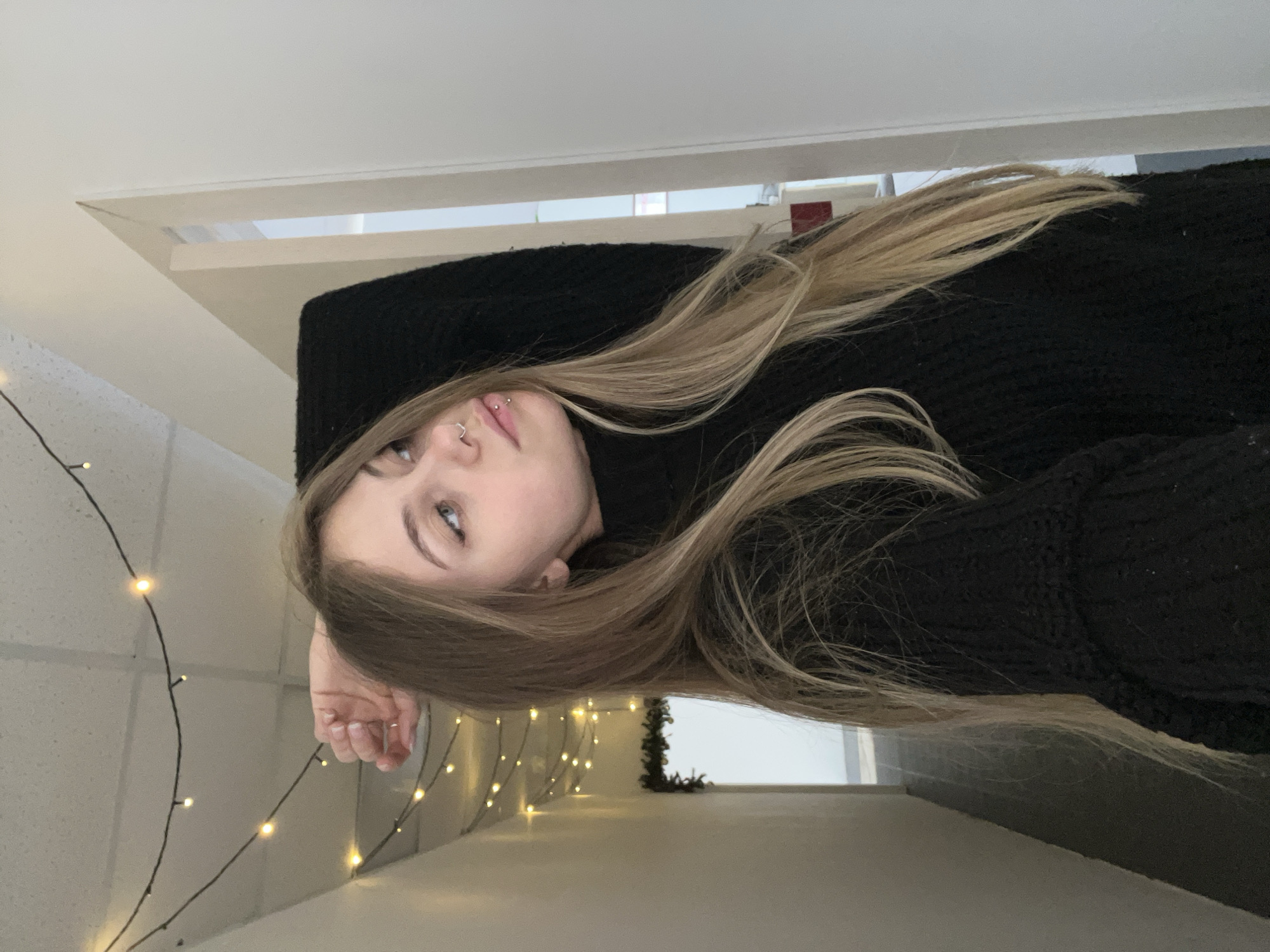 The image shows a young woman with light blonde hair, wearing a black sweater. She is lying down, with her head tilted to the side. Her face is visible, showing her eyes and nose, which has a piercing. Her left hand is visible near the edge of the frame. In the background, there is a wall decorated with string lights and a window. There's no evidence to suggest this photo was taken in Фоки, Russia.