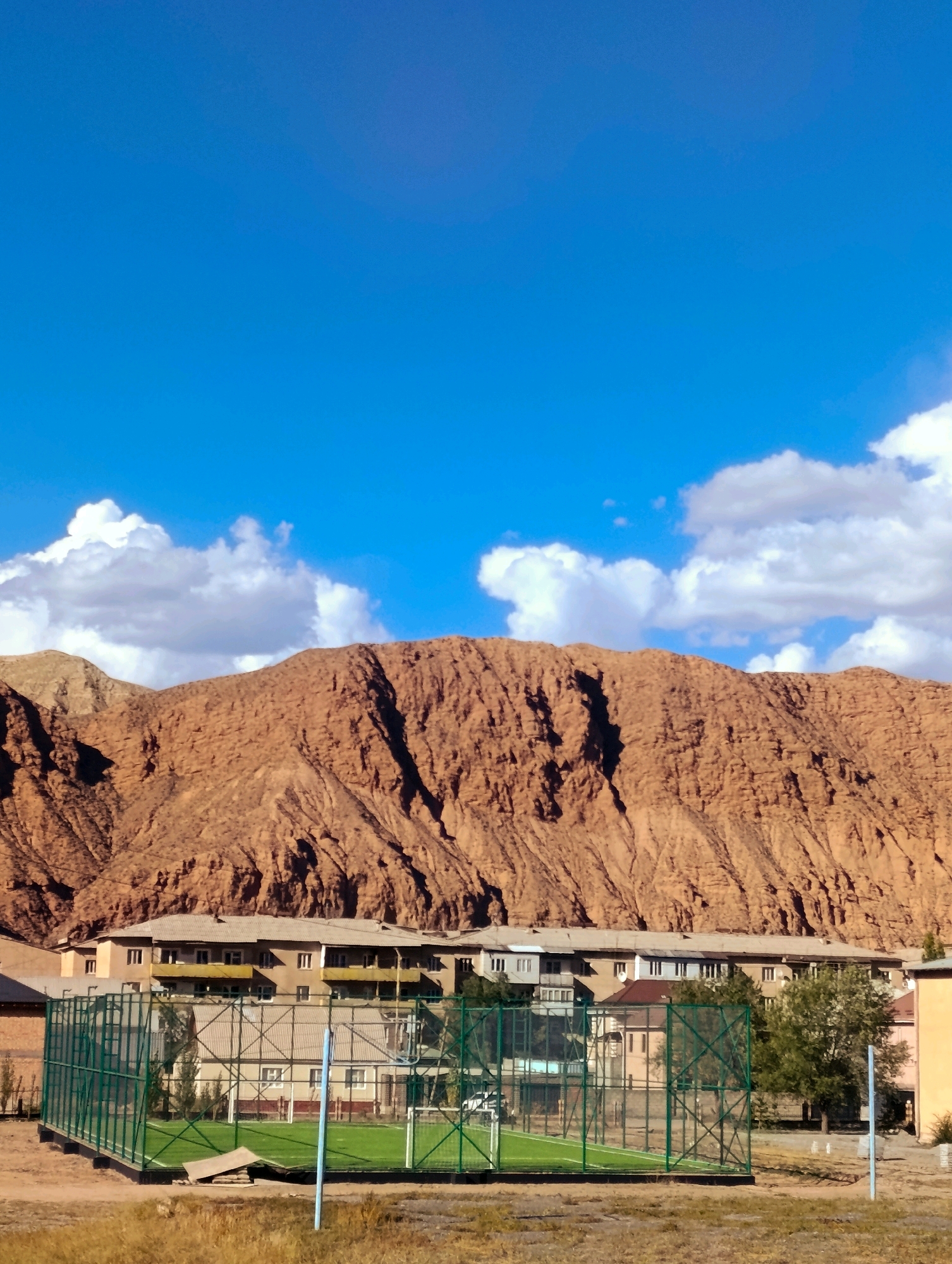 The image shows a view of Naryn City, Kyrgyzstan, with a mountain range in the background. The foreground features a green fenced sports court, possibly a soccer or basketball court, with artificial turf. Behind the sports court are several multi-story buildings, appearing to be residential apartments. The buildings are of a neutral color palette with some balconies. Further behind the buildings, a large, rugged, red-brown mountain rises, dominating the middle ground of the image. The sky is a vibrant blue with scattered white clouds. The overall impression is of a somewhat arid or mountainous urban environment under a clear sky. There are no people visible in the image.