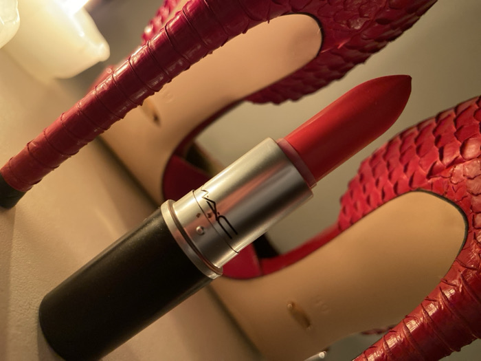 A close-up shot features a MAC lipstick with a red hue, prominently displayed against a backdrop of red snakeskin stilettos. The lipstick's silver casing and black base are clearly visible. The heels, with their textured material, add depth to the composition. A soft light source illuminates the scene, highlighting the textures of the lipstick and shoes, as well as the color contrast between the objects.