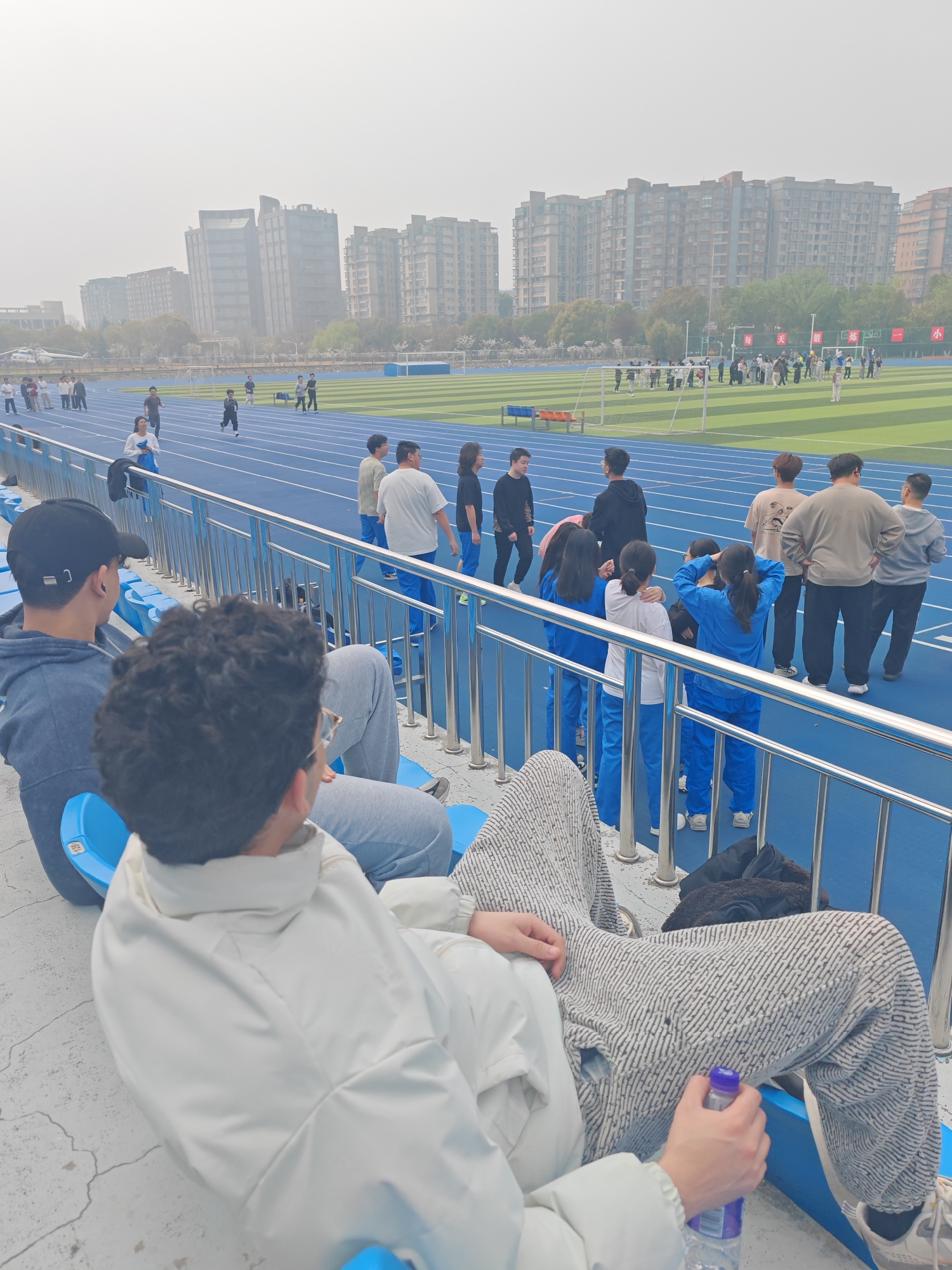 The image depicts a scene at a track and field stadium in Jiangning District, China. The stadium features a blue running track, with groups of people standing and walking on it. The scene is viewed from the stadium seating, where several people are sitting. One person, wearing a white jacket and patterned pants, is prominently positioned in the foreground holding a water bottle. The sky is overcast with a visible haze obscuring buildings in the background.