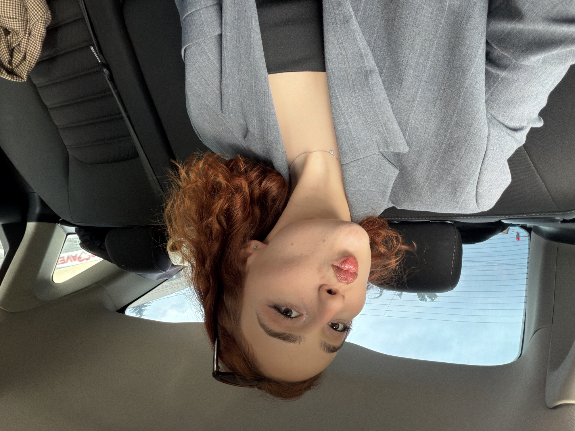 A reddish-haired woman is seated in the back of a car, facing the camera, her head upside down. She wears a gray pinstripe blazer over a black top, and a delicate bracelet is visible on her neck. The car's interior includes black leather seats and a dark headrest. Through the rear window, a bright sky and treetops can be seen, with a partial view of a sign that includes the letters "AVE," which suggests she might be in a parking lot or driving by a business with a sign including these letters. Considering the information provided, it's not possible to accurately tell the location in the city of Tbilisi, Georgia.