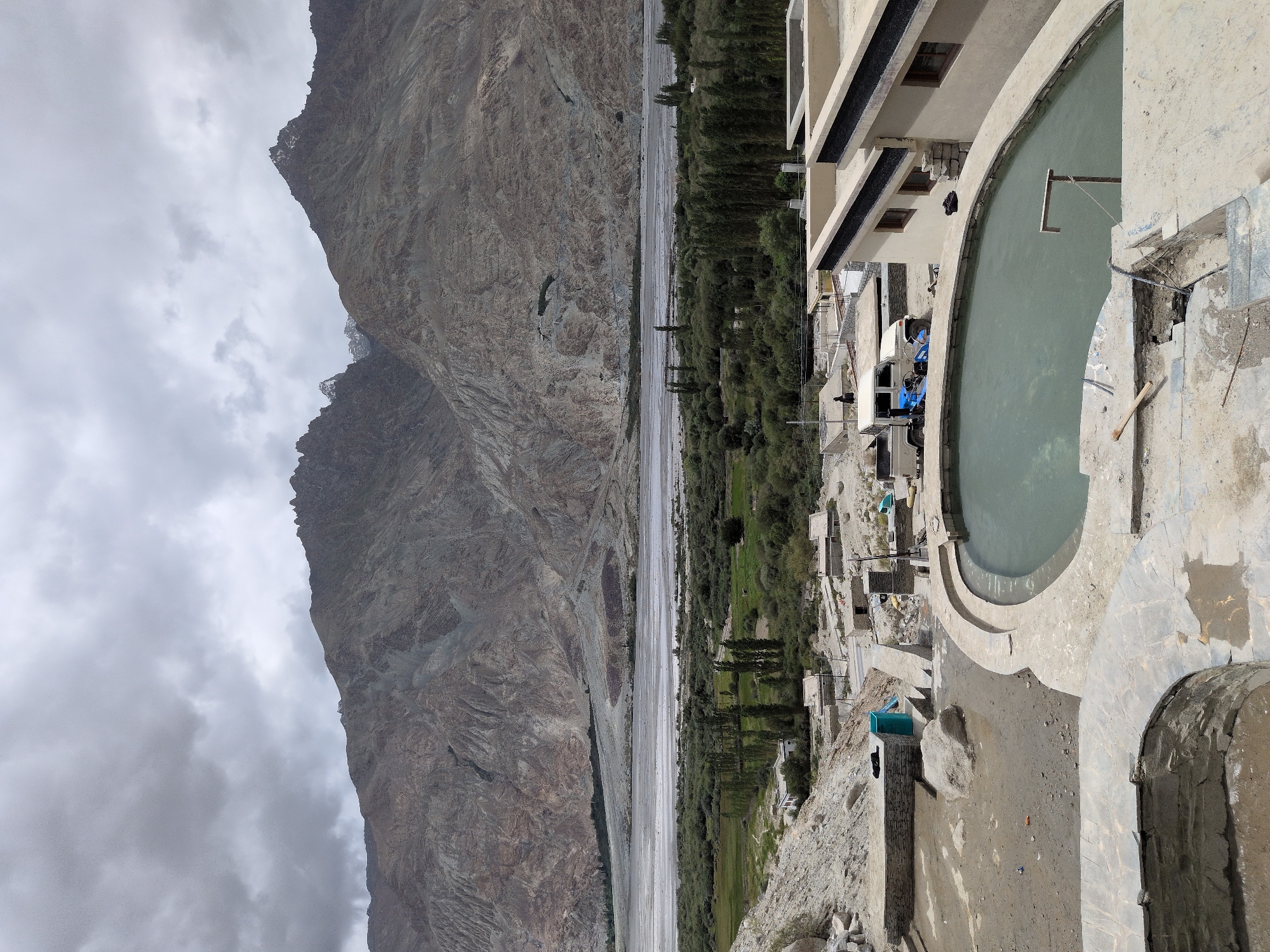 The image depicts an outdoor hot spring in Panamik, India, nestled amidst a rugged, mountainous landscape under a cloudy sky. In the foreground, a kidney-shaped pool filled with cloudy, greenish water is the central feature, surrounded by concrete and stone construction that appears to be in a state of partial completion or repair. Tools, including a wooden-handled object resembling a spade or hoe, are visible on the concrete.
Beyond the hot spring, a collection of small, traditional buildings with flat roofs are scattered across the terrain. A light-colored utility vehicle and a blue piece of equipment are parked near one of these structures. Further in the background, a wide, arid riverbed stretches out, bordered by lush green vegetation, including a line of tall trees. The middle ground transitions to a verdant valley with more trees and small clusters of buildings.
Dominating the background are immense, imposing mountains with jagged peaks, their rocky surfaces textured and eroded by time. The overall impression is one of a remote, natural setting enhanced by man-made structures. The cloudy sky casts a diffused light over the scene, contributing to a subdued color palette. No individuals are clearly identifiable as actively participating in any activity within the frame.
