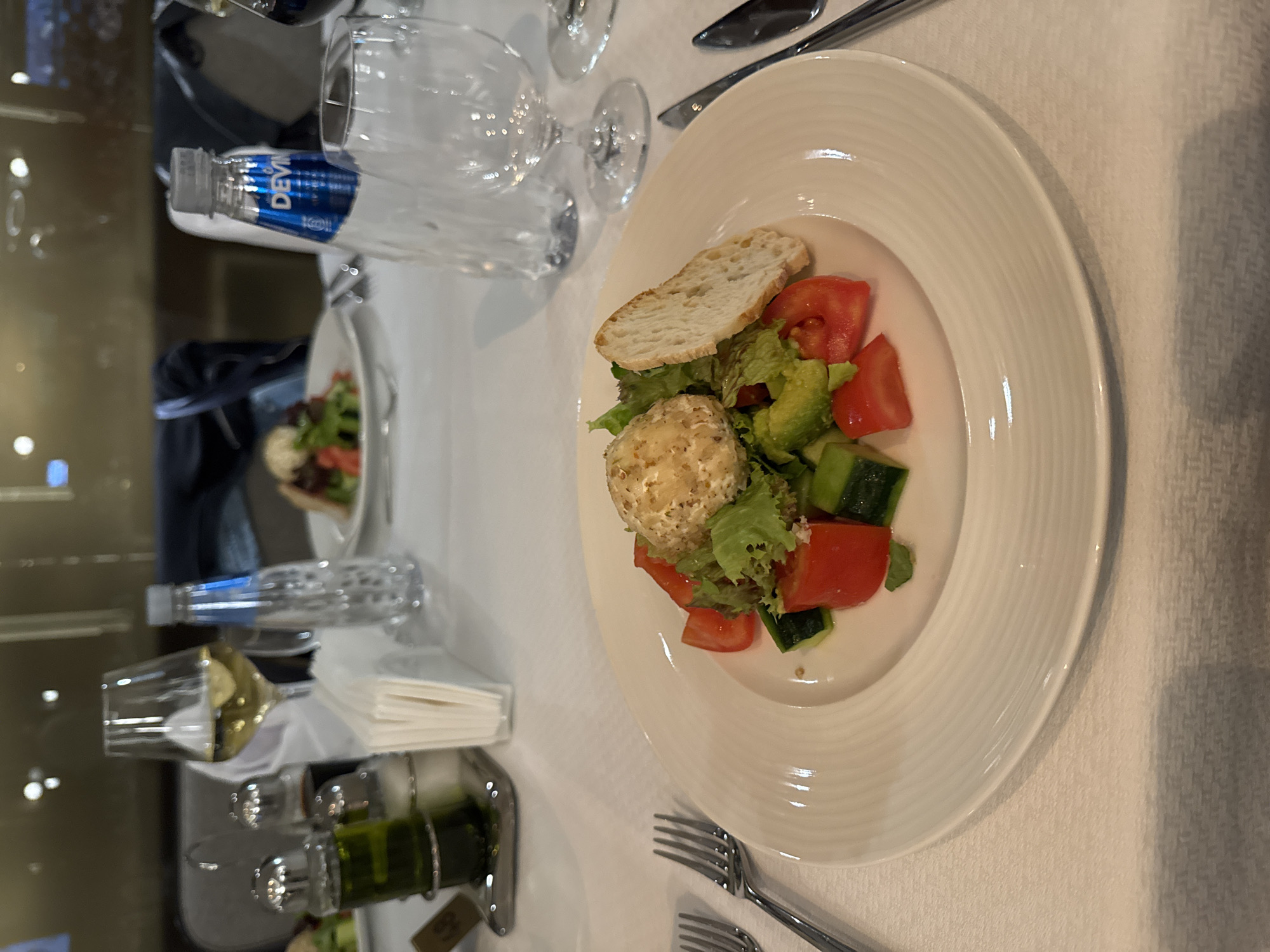 The image shows a formal dining table setting, likely in a restaurant. The scene is well-lit. A plate of salad is prominently displayed. The salad contains mixed greens, tomatoes, cucumbers, avocado, and a bread slice, topped with a ball of cheese. There's a bottle of water, a wine glass with white wine, cutlery, and a napkin holder. A second plate with salad is visible in the background.
The location cannot be definitively determined as Varna, Bulgaria, without additional context or clues.