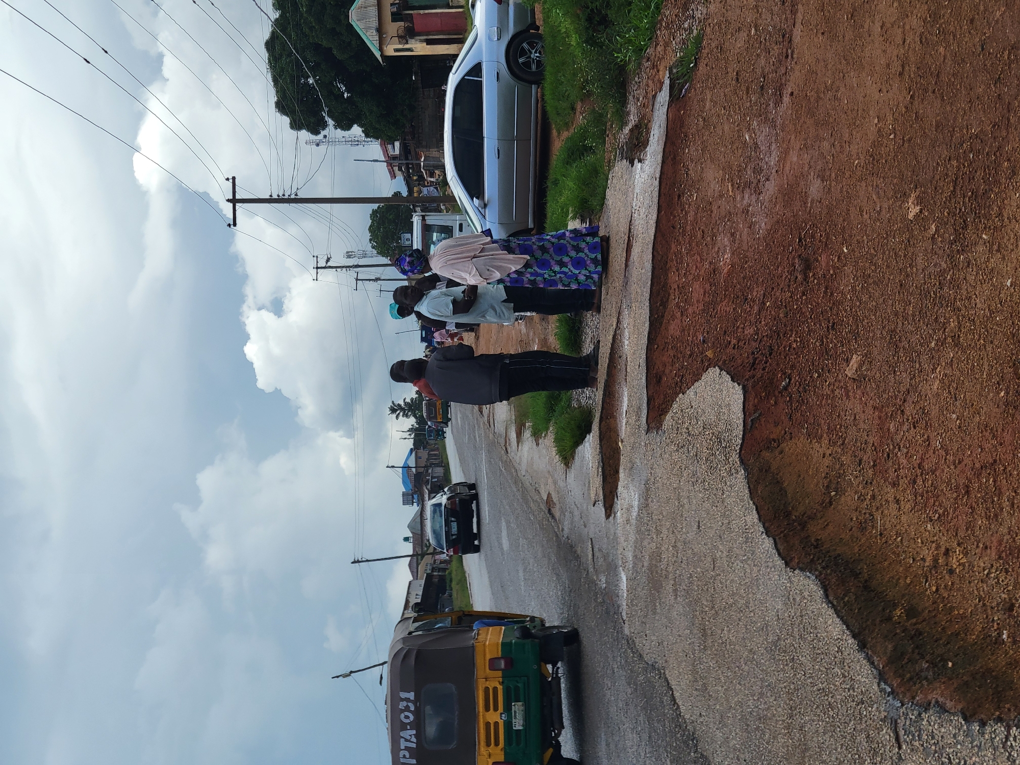 The image shows a street scene in Jos, Nigeria, with a damaged asphalt road in the foreground. A tuk-tuk is parked on the left side of the road, facing away from the camera. Several people are standing on the right side of the road, engaged in conversation. A silver sedan is parked behind them. In the background, there are buildings, trees, and power lines, with a cloudy sky overhead. The damaged road suggests potential infrastructure issues in the area.