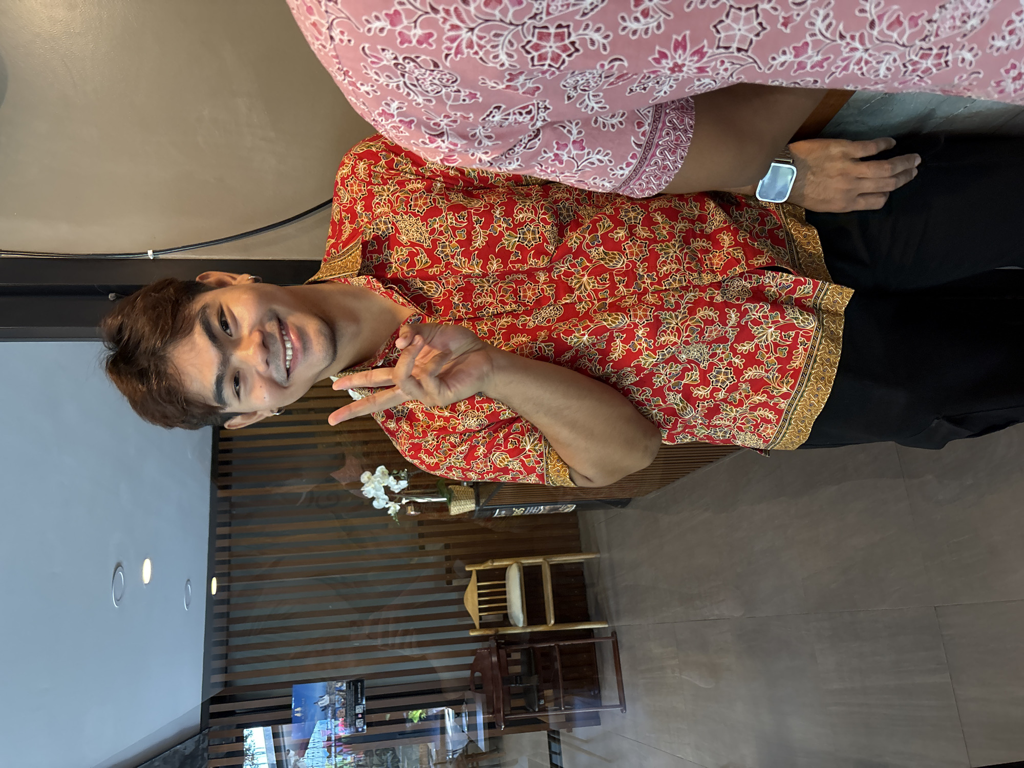 A man wearing a red patterned shirt and black pants is posing for a photo. He is smiling and holding up two fingers in a peace sign. Another person wearing a pink patterned shirt is partially visible above him. The photo appears to be taken indoors in a cafe or restaurant in บ้านบางรักษ์, Thailand, as indicated by the wall decorations and furniture in the background.