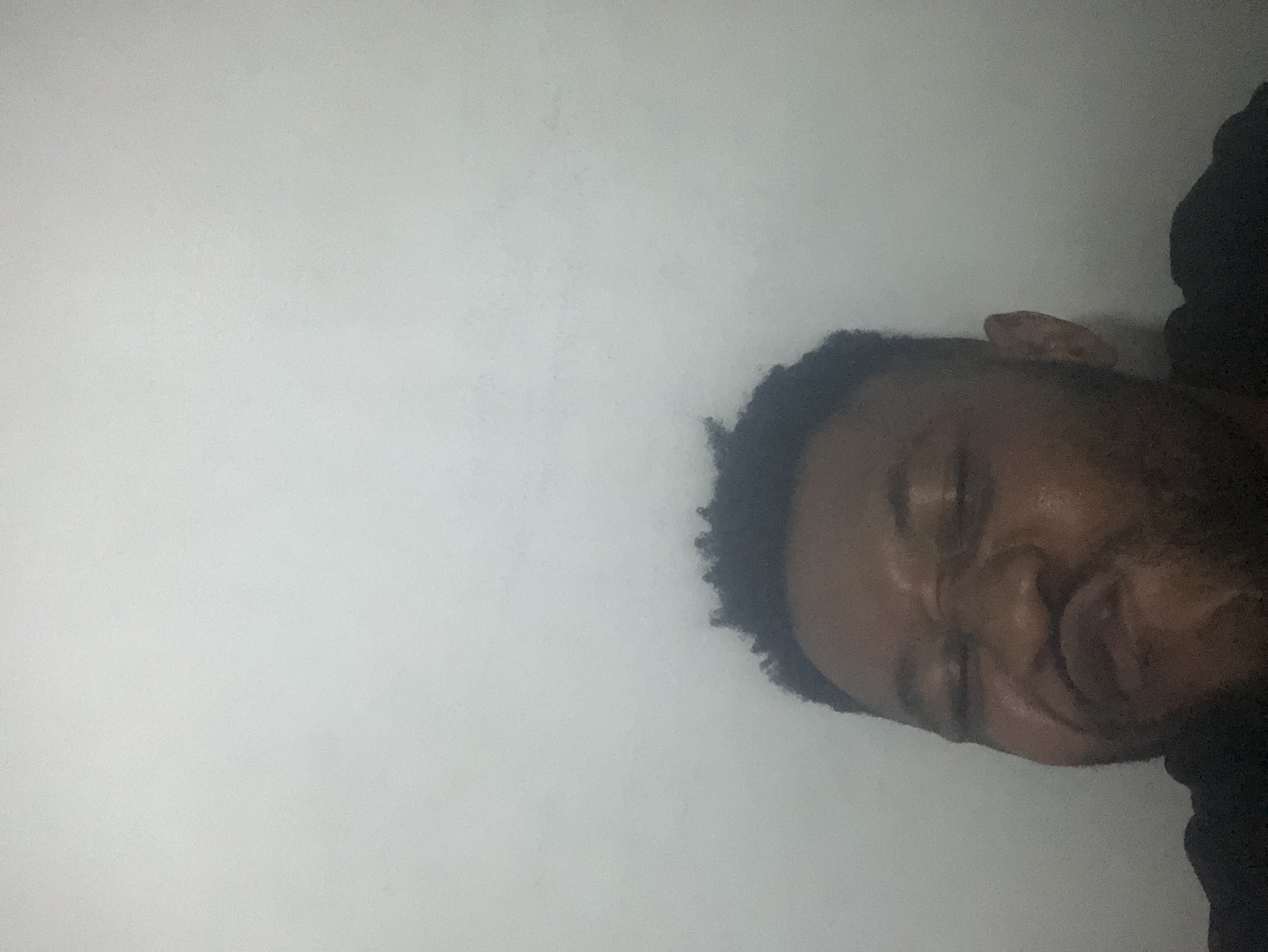 The media file displays a close-up portrait of a dark-skinned man, oriented sideways with his head positioned on the right side of the frame, tilted approximately 90 degrees counter-clockwise from an upright posture. He has short, dark, curly hair. His eyes are partially closed or squinted, and his mouth forms a subtle smile or smirk, conveying a relaxed or content expression. Part of his right ear is visible on the upper right side of his head. He appears to be looking towards the right side of the image as presented. The background is a plain, light grey or off-white surface, likely a wall, occupying the majority of the left and top areas of the image. The lighting is soft, creating gentle shadows on his face without stark contrasts. The overall image possesses a slightly grainy texture. No visible text is present. The man is wearing dark clothing, potentially a black shirt, which is visible as a dark shape along the bottom right edge of the frame. The time of day and specific location cues are not discernible beyond an indoor or generic setting.