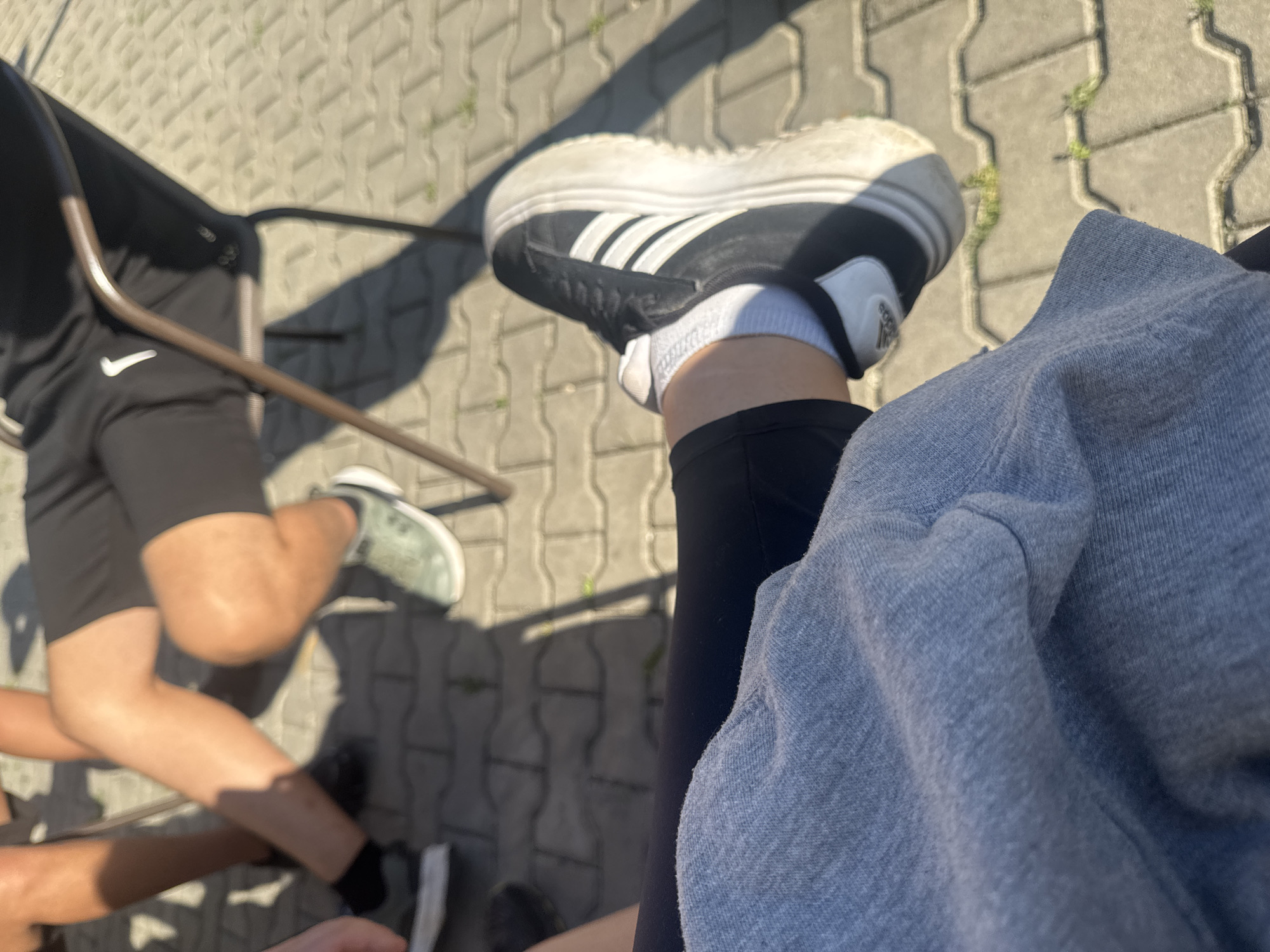 The image shows an outdoor scene with a group of people sitting on folding chairs. The ground is paved with grey interlocking blocks.
One person in the foreground is wearing black pants, white socks, and black and white Adidas sneakers. A light blue sweatshirt is draped over their lap. To the left, another person is wearing black shorts with a white Nike logo and light green sneakers. There is another person in the bottom left wearing black socks.