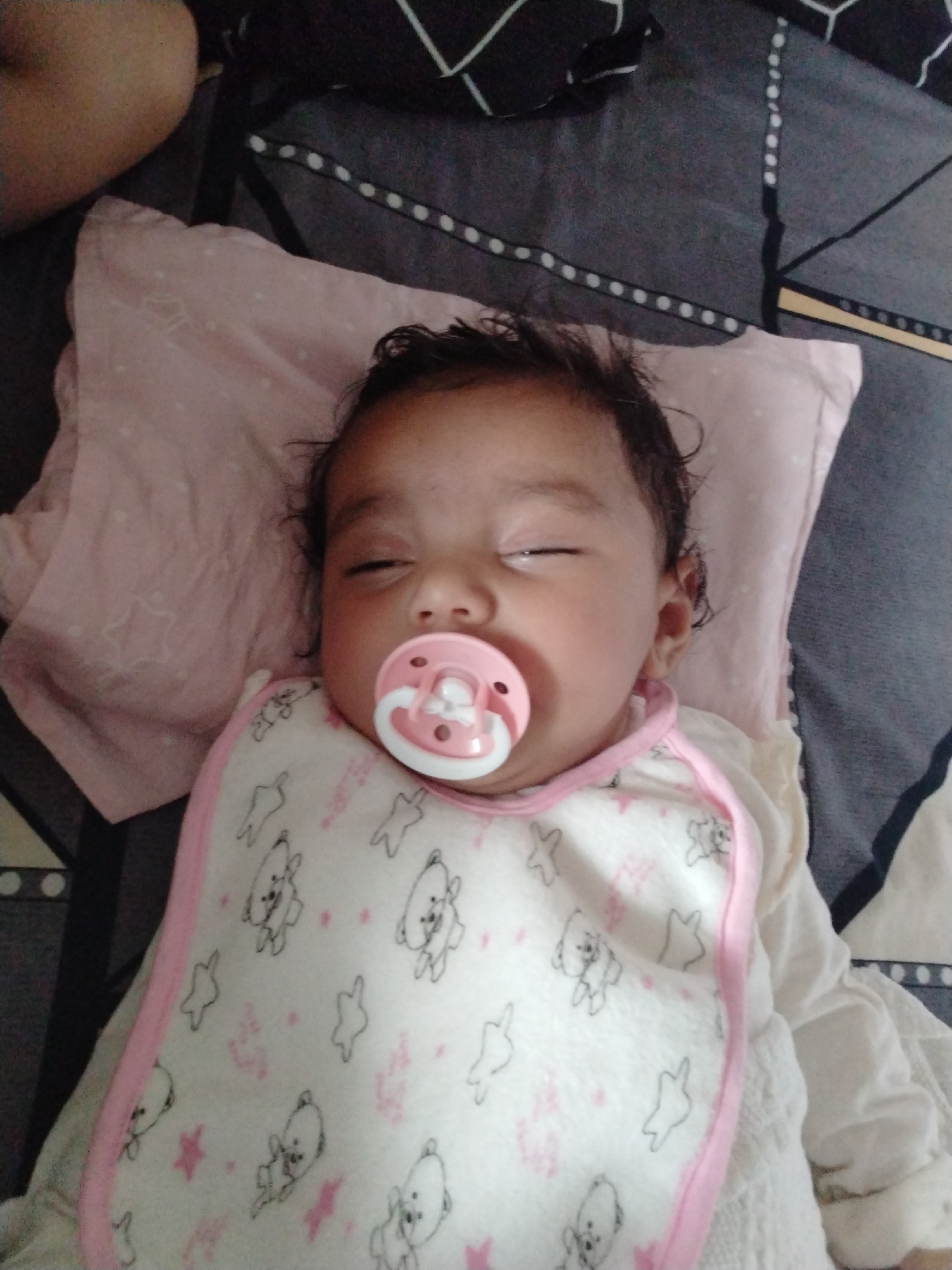 A close-up, top-down shot depicts a sleeping baby with a pacifier in their mouth. The baby is lying on a light pink pillow on what appears to be a patterned bedsheet. The baby is wearing a white bib with a pink trim, adorned with illustrations of bears and stars. The location is Unknown, Unknown.