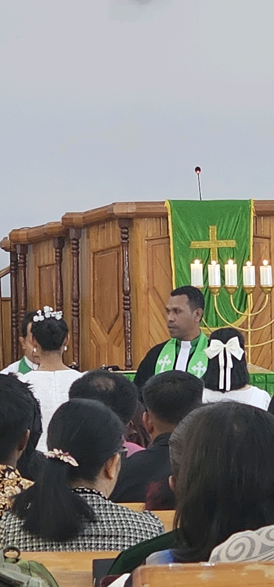 The image depicts a scene inside a church in Puncak Jaya, Indonesia. In the foreground, the backs of several attendees are visible, seated on wooden pews. Some individuals have dark hair, and one person in the center has dark hair tied back with a decorative clip. Moving further into the frame, a person wearing a white dress and a floral hair accessory is partially visible from the back. To their right, a man in clerical attire, including a black robe with a green stole featuring a white cross, stands facing towards the right of the image. Behind him, a green altar cloth with a cross is draped over a wooden structure, and five lit candles are arranged on a candelabra. The church interior features wooden paneling and decorative elements on the walls and furniture. The overall lighting suggests an indoor setting, possibly during a service or ceremony.