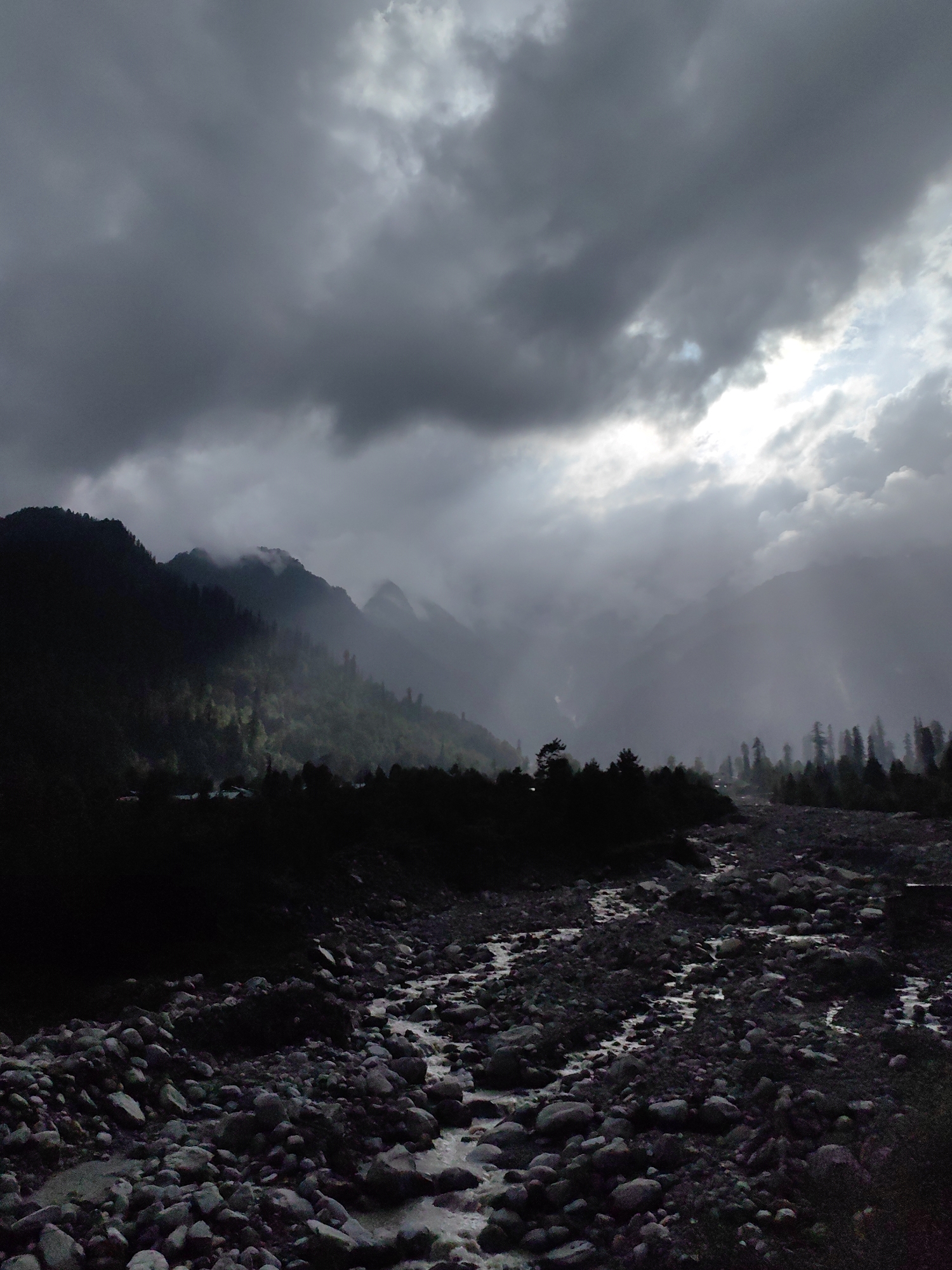 The image depicts a natural landscape with a rocky riverbed in the foreground. The riverbed is filled with various sizes of stones and pebbles, with water flowing through channels between them. In the mid-ground, a dark silhouette of trees and hills is visible. The background features a mountain range shrouded in mist and dramatic, dark clouds. There is a visible shaft of light breaking through the clouds, illuminating some of the mist. The scene appears to be in a natural, unpopulated area. The location is Palchan, India, which is known for its mountainous terrain and natural beauty.